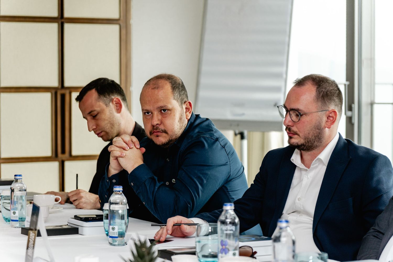 Three businessmen sitting at a conference table during a professional meeting with water bottles and documents.