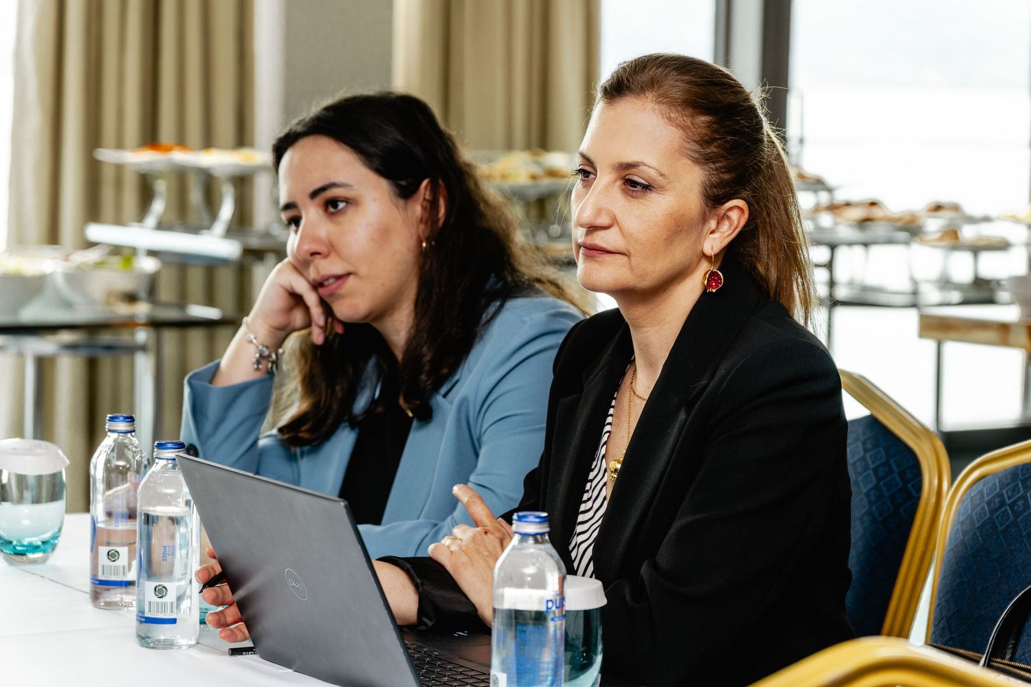 Two professional women sitting at a conference table with laptops and water bottles during a business meeting.