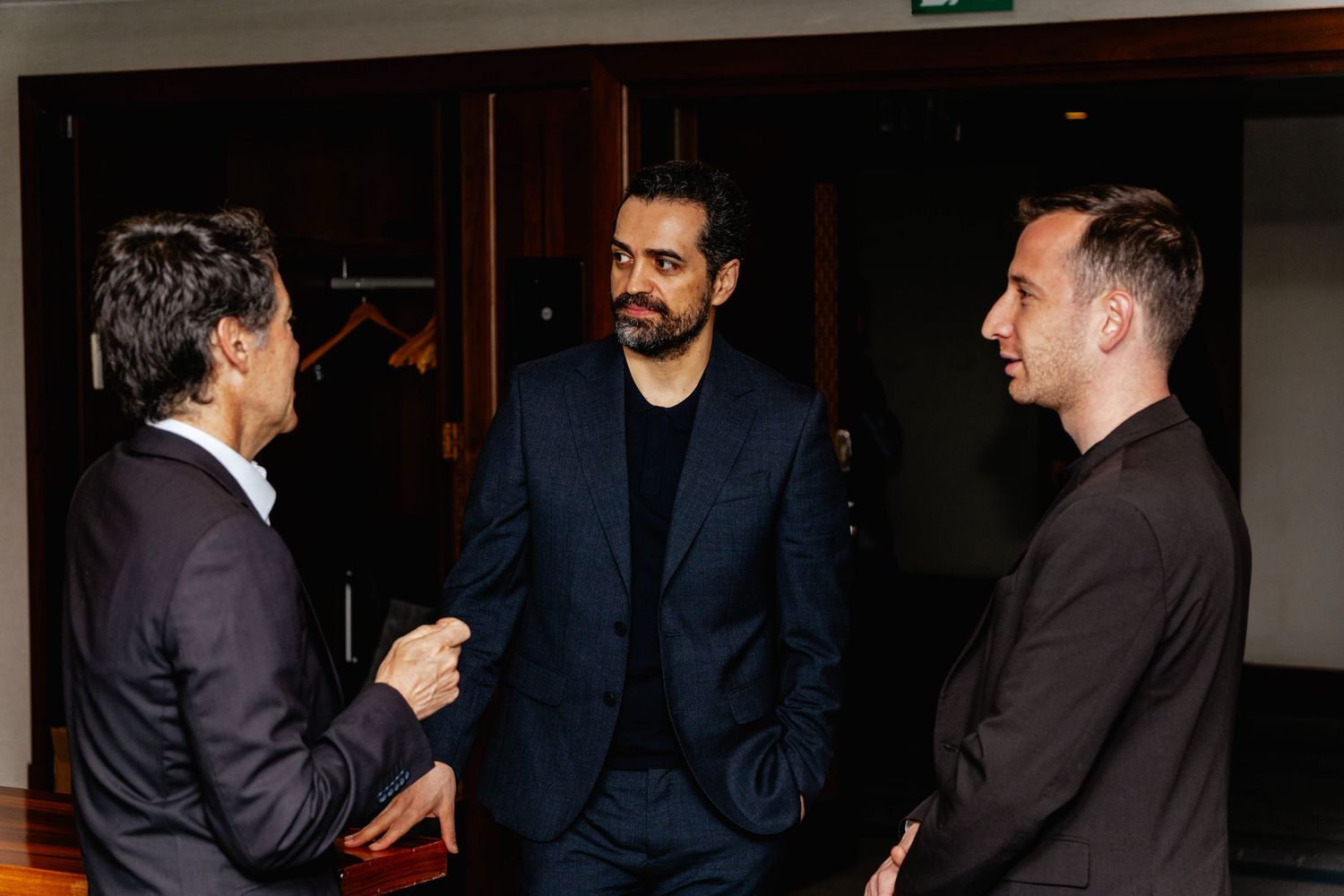 Three men in formal attire engaged in conversation at an elegant indoor networking event.