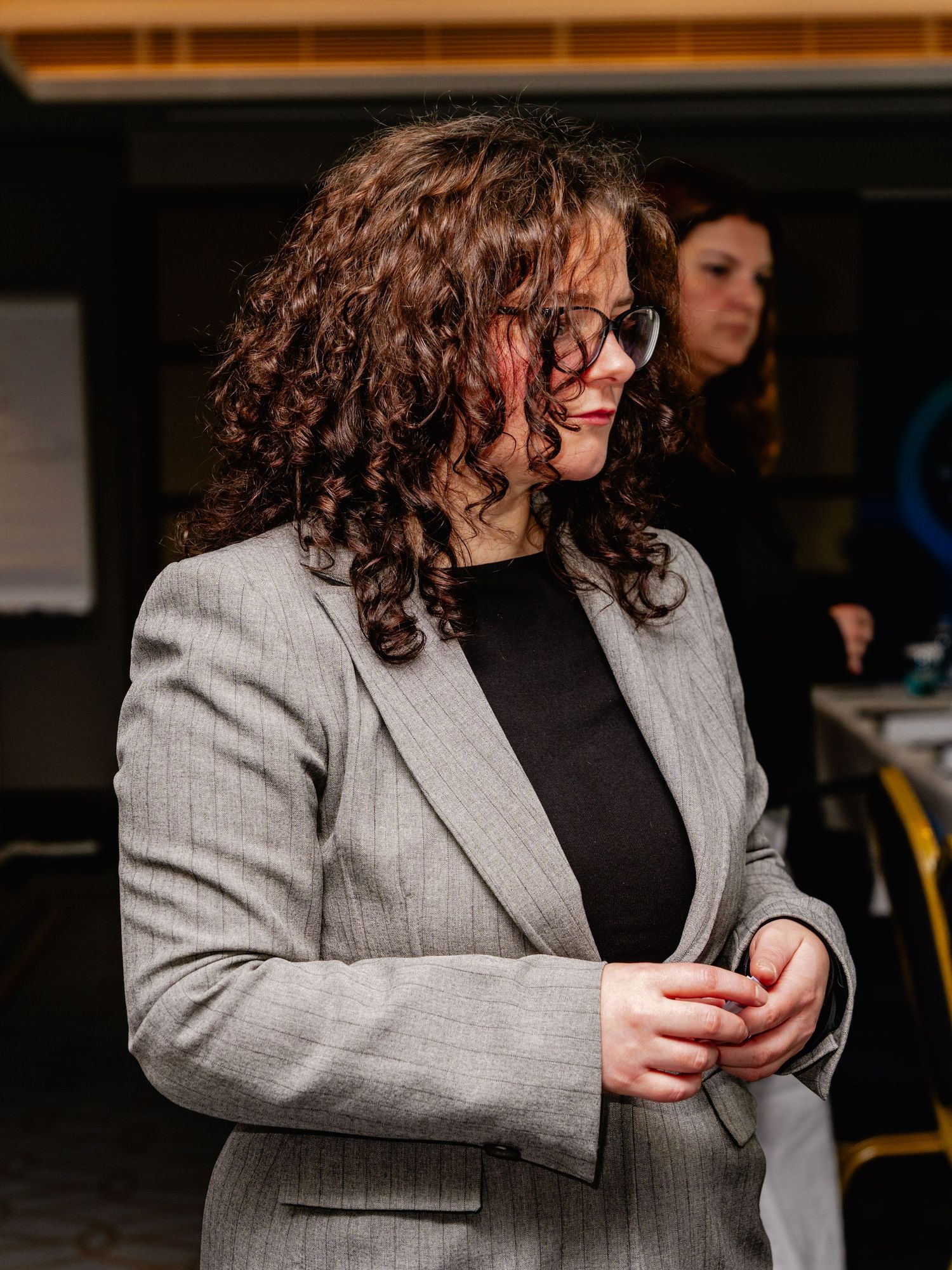 Woman with curly hair and glasses wearing a gray blazer over black top stands at an indoor professional event.