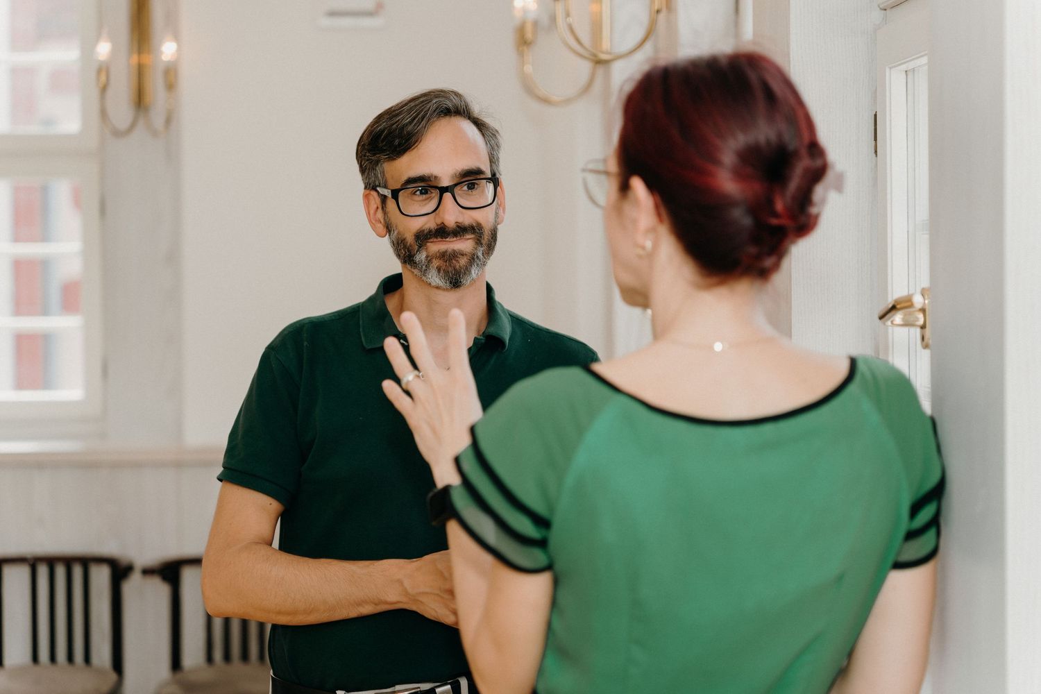 Man with glasses and beard in green polo talks with red-haired woman in green top in bright room.