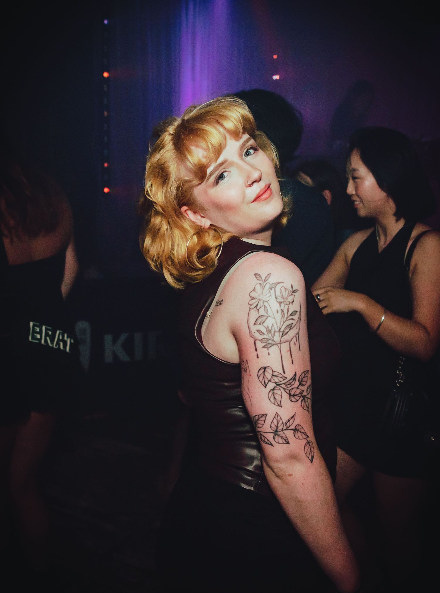 People dancing in a dimly lit nightclub with colorful purple lighting casting a moody atmospheric glow.