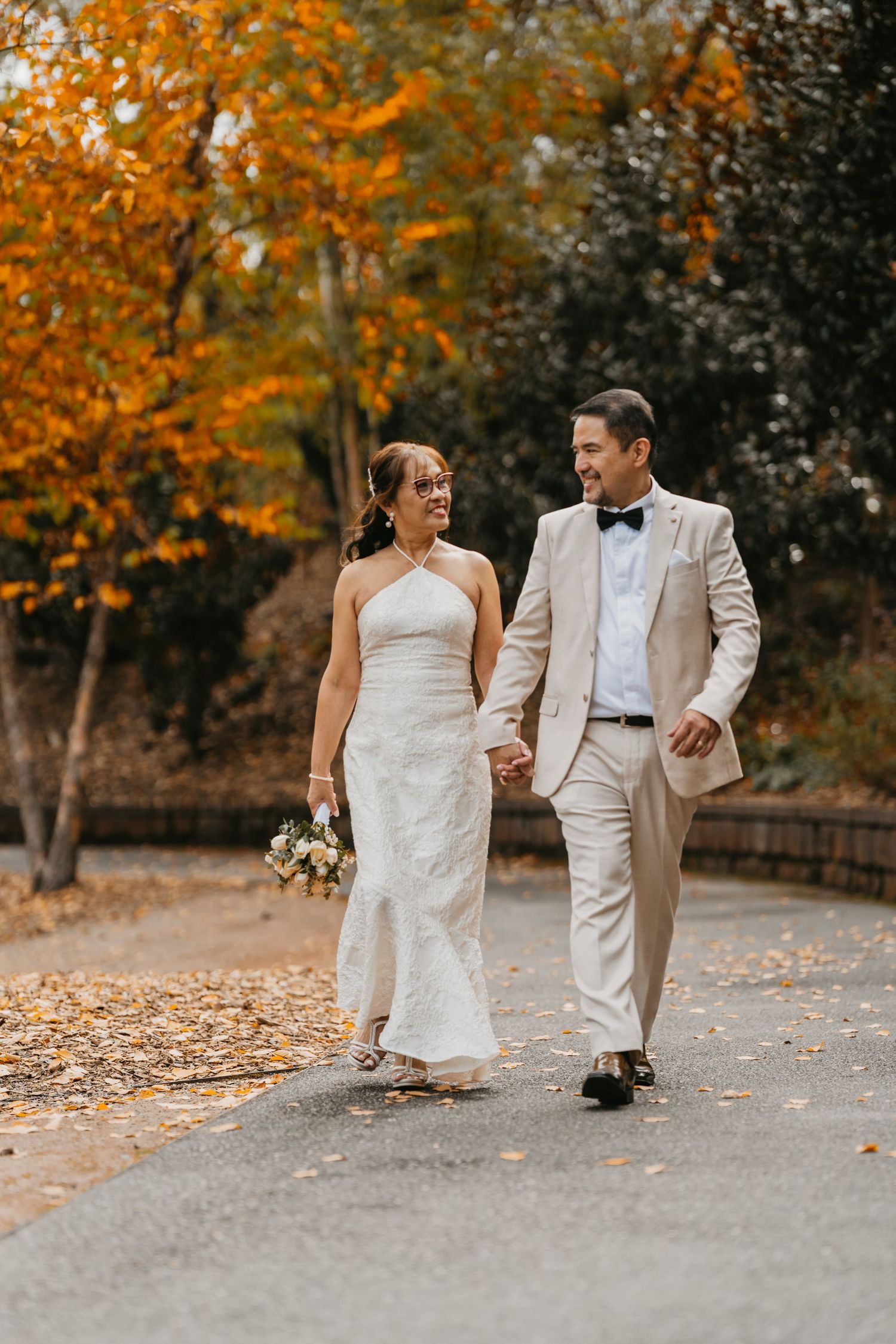 A wedding couple walks and laughs together on a path surrounded by vibrant orange autumn foliage.
