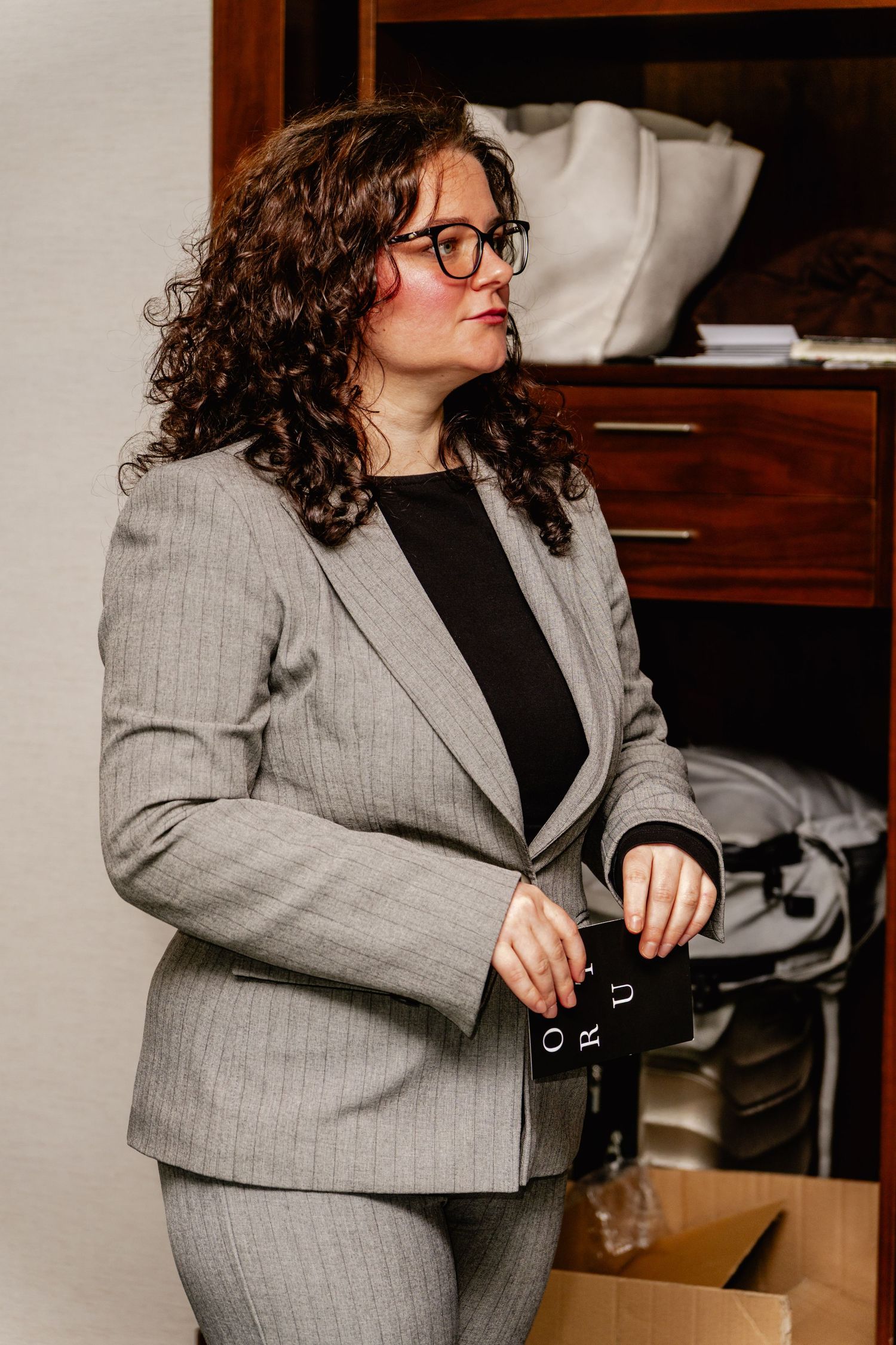 Woman in gray suit with glasses holding a black item, standing near a wooden cabinet in an office setting.