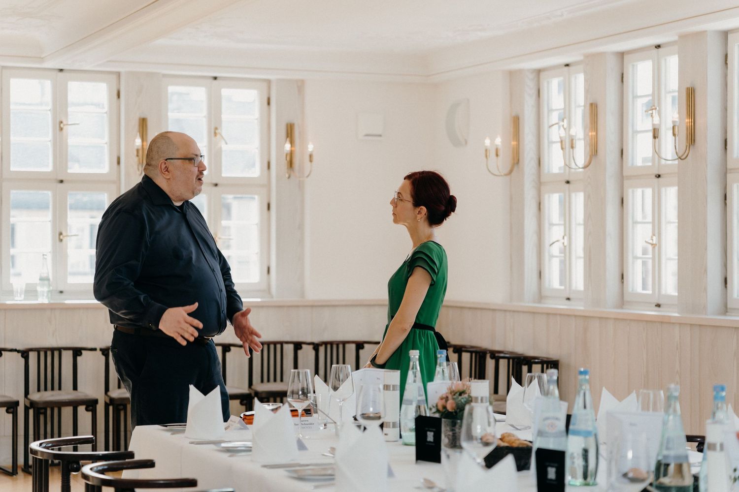 A man in black and woman in green dress converse beside an elegantly set dining table in a bright event room.