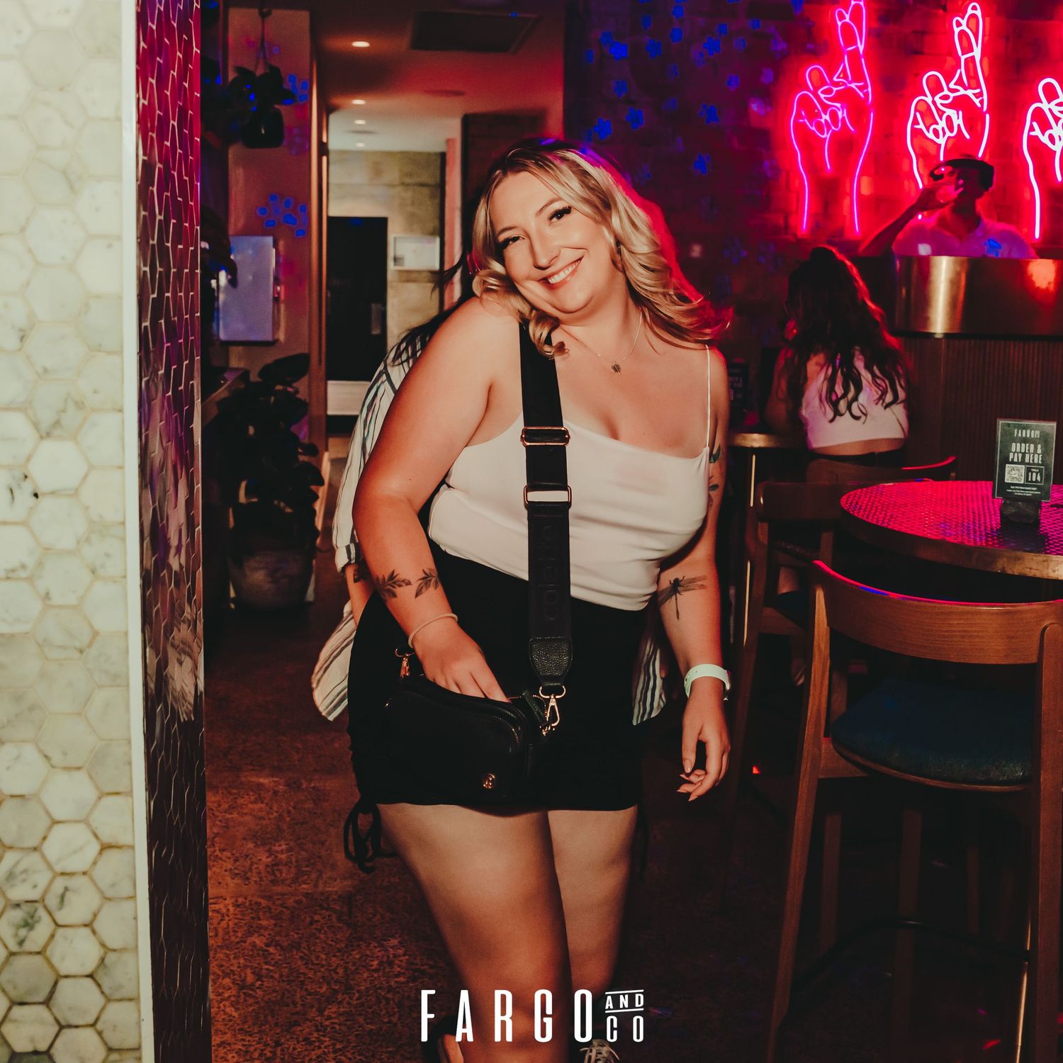 Woman in black shorts and beige top poses against dark interior with glowing neon red lights and bar decor in background.