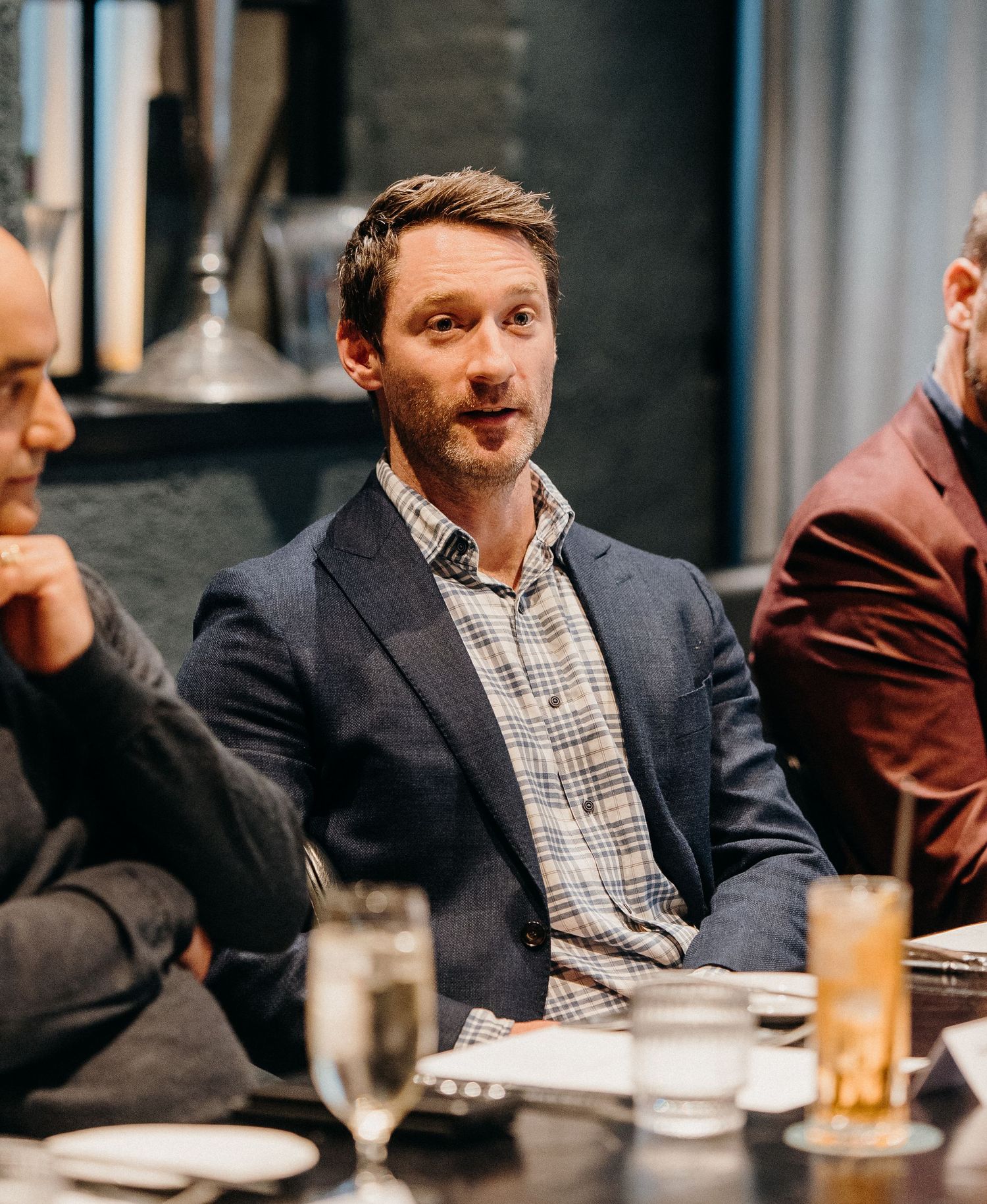 A person in a suit jacket and checkered shirt sits at a table with drinks during a social gathering.