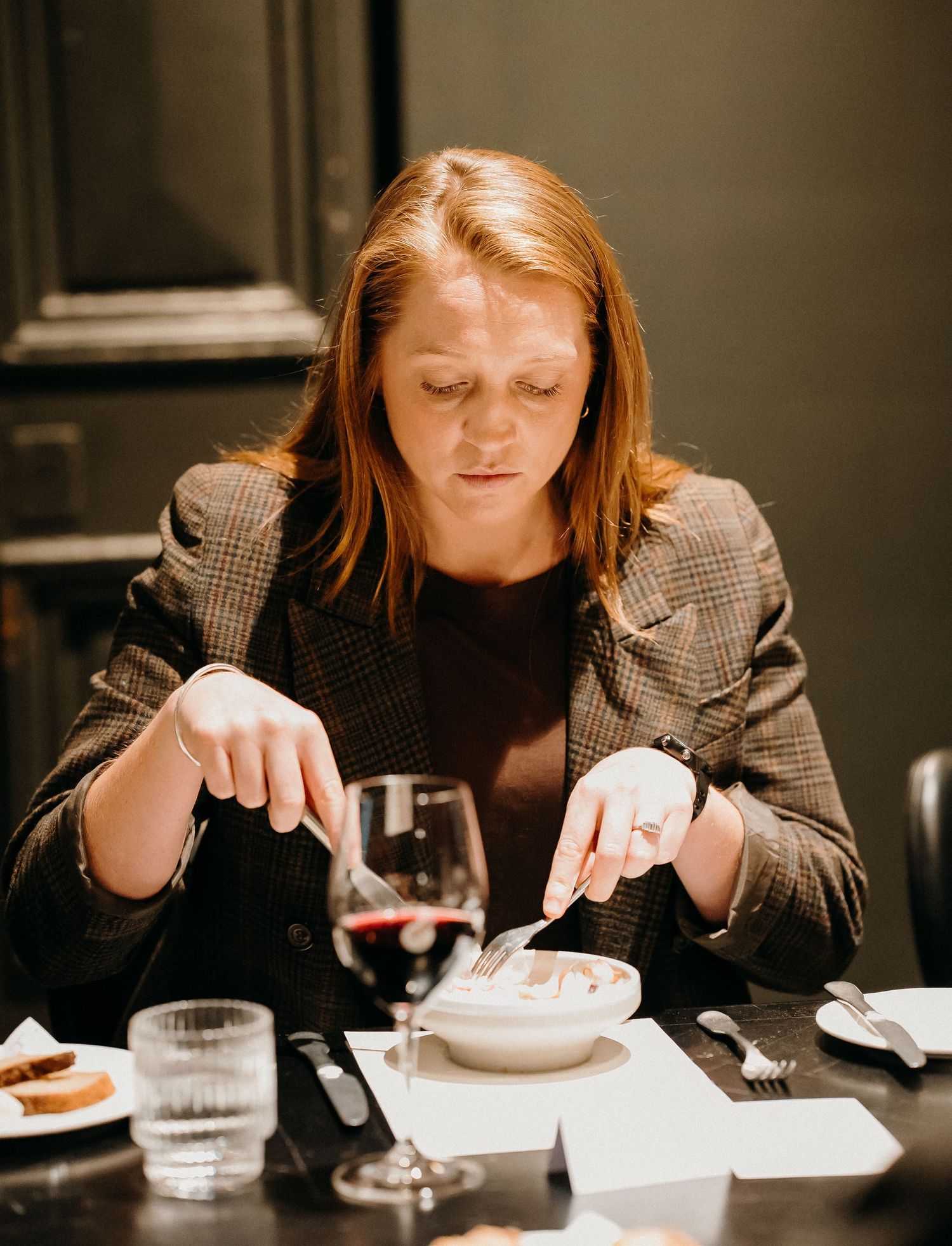 A person in a brown blazer sits at a dining table eating and drinking from multiple dishes in a dimly lit setting.
