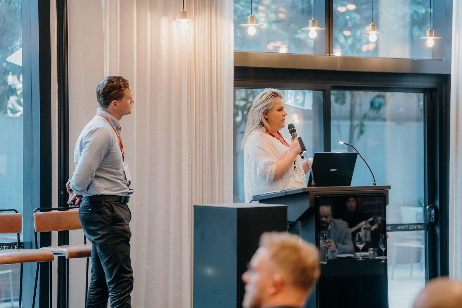A speaker stands at a podium delivering a presentation in a modern office space with pendant lighting.
