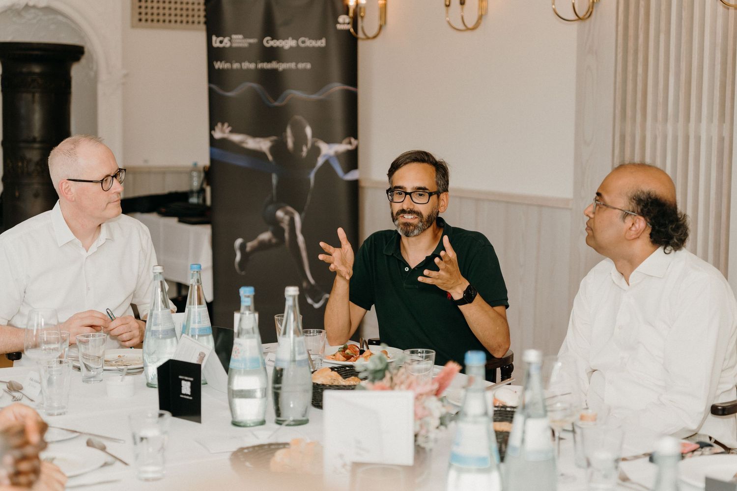 Three businessmen engaged in discussion at a Google Cloud event dinner table with water bottles and food.