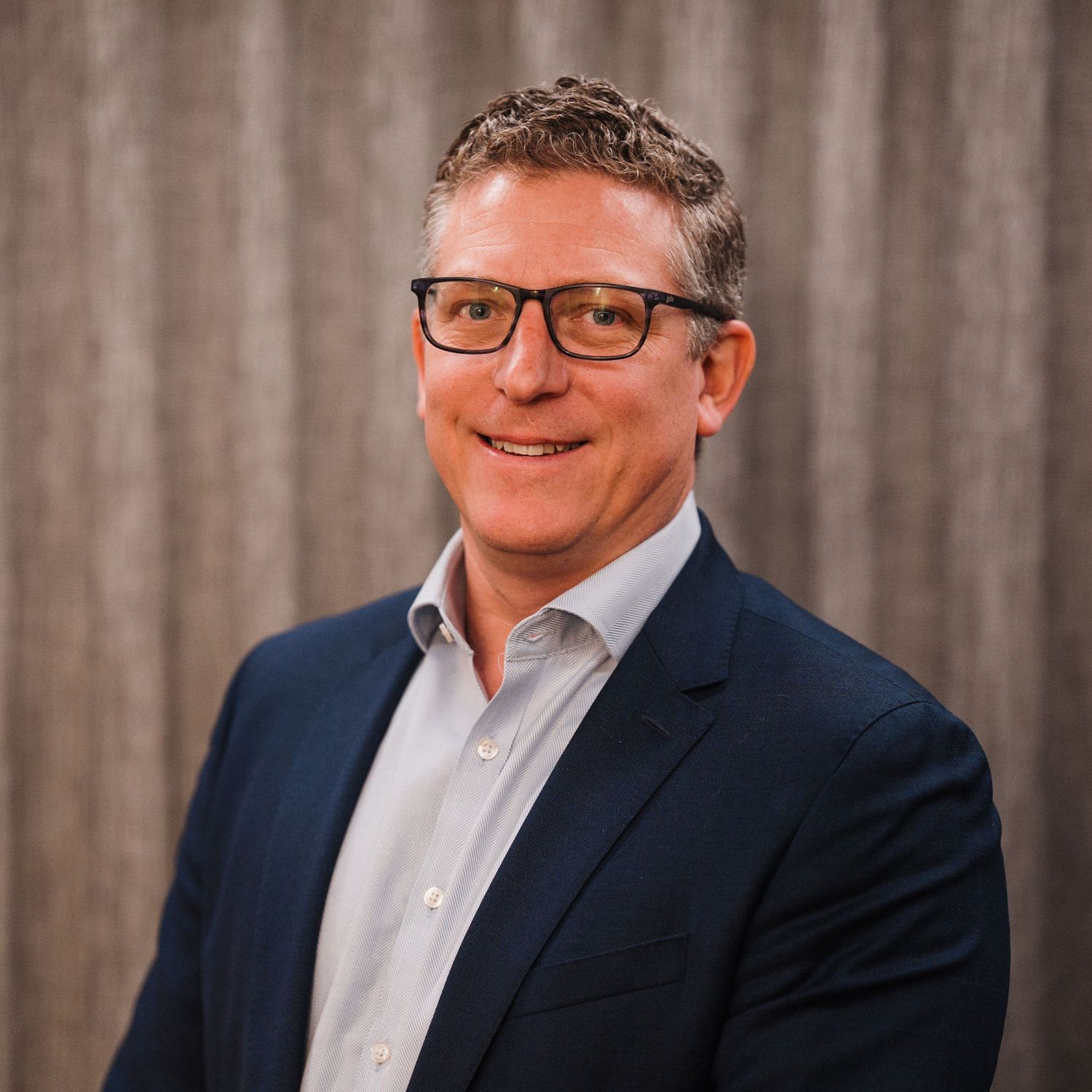 Professional headshot portrait of a person wearing glasses and a business suit against a gray background.