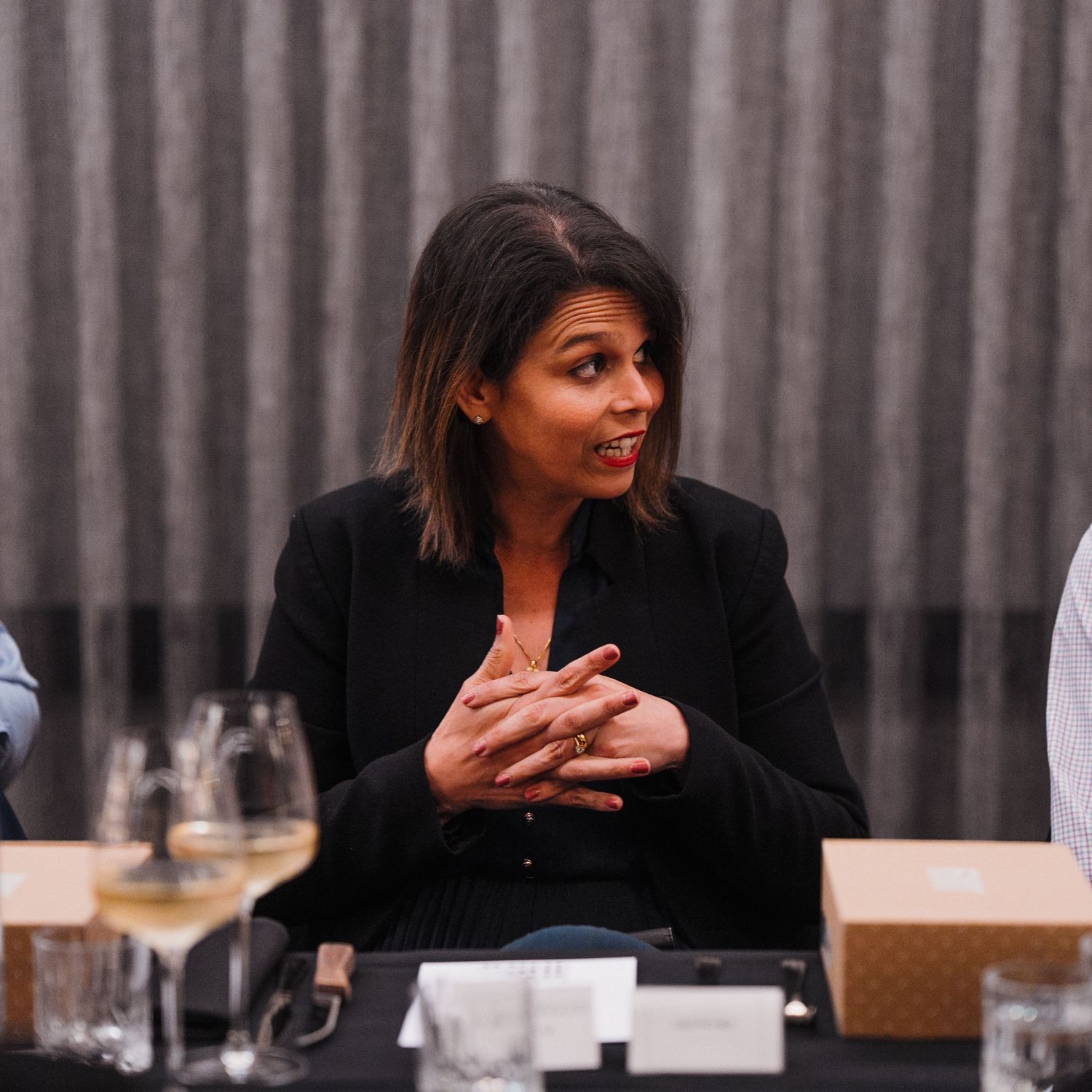 Professional woman in black blazer gestures animatedly while speaking at business dinner meeting.