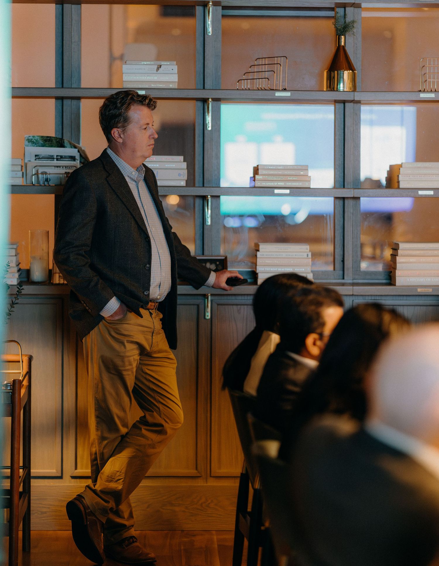 A person in business attire stands near illuminated shelving units displaying books and wine bottles in an office setting.