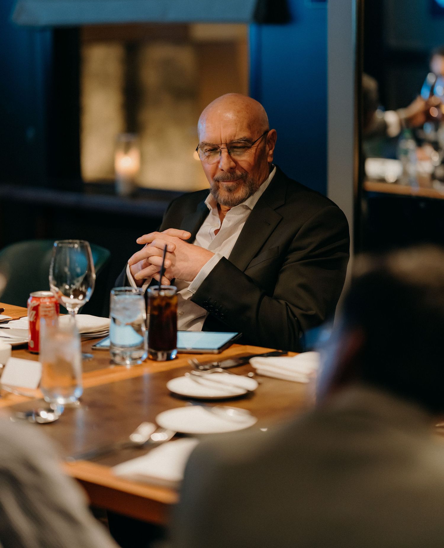 A person in a black suit sits at a restaurant table enjoying a meal and drinks.