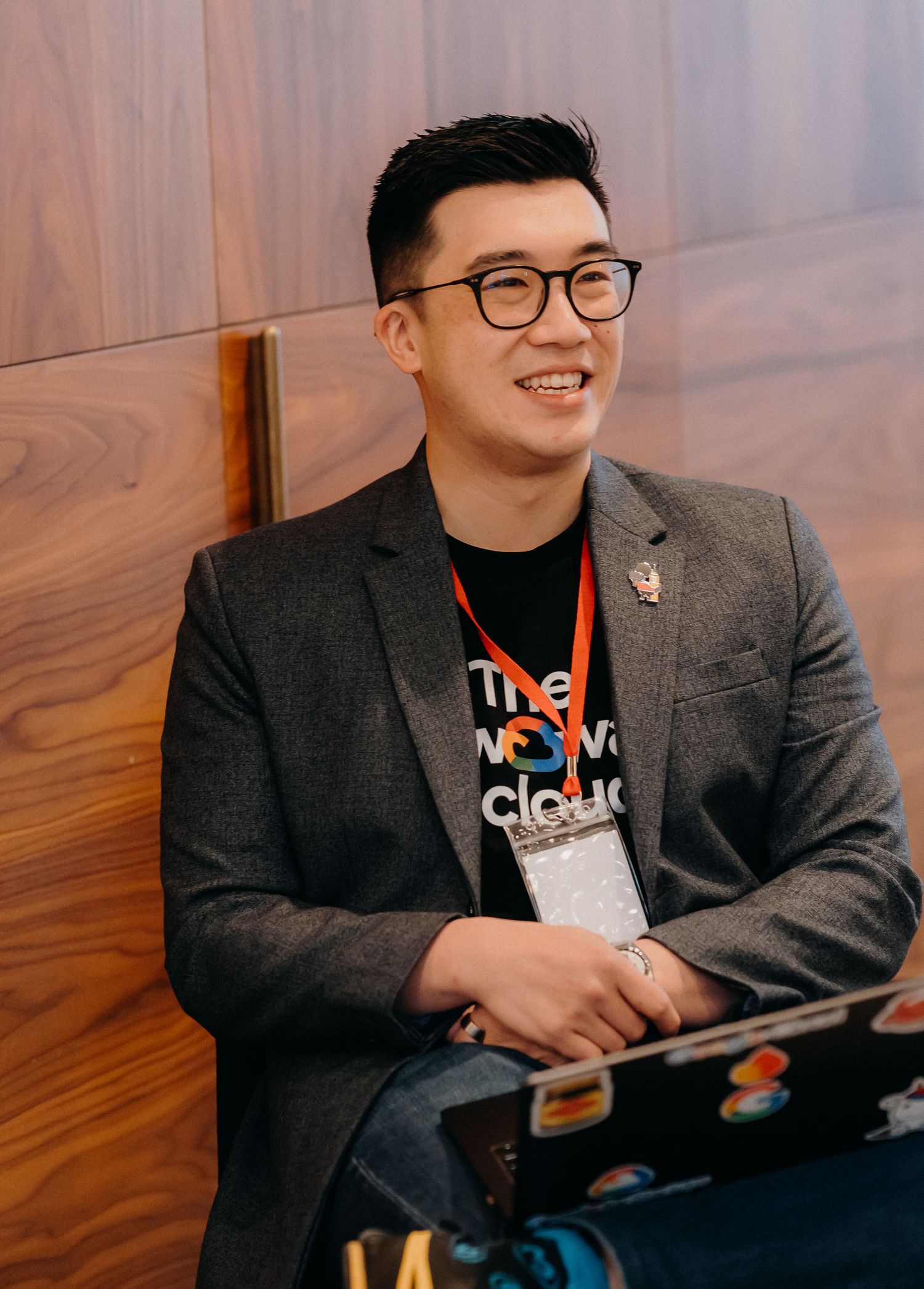 A person in a dark blazer and glasses sits casually in front of a wooden wall while smiling and gesturing.
