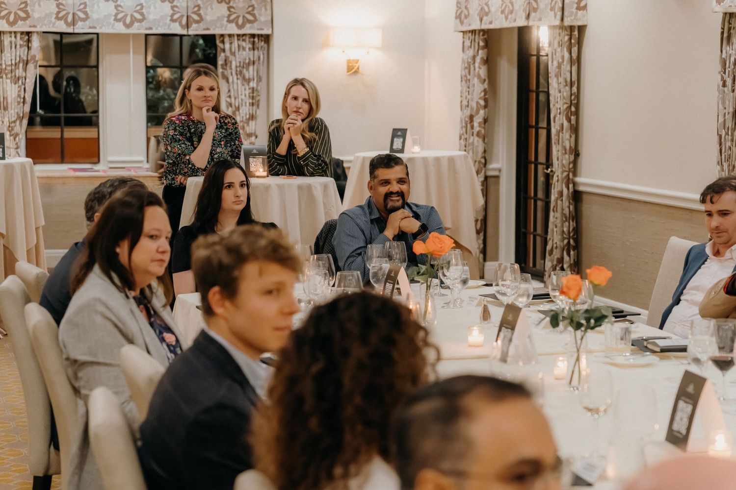 Wedding guests seated at round tables during an elegant indoor reception with white tablecloths and floral centerpieces.
