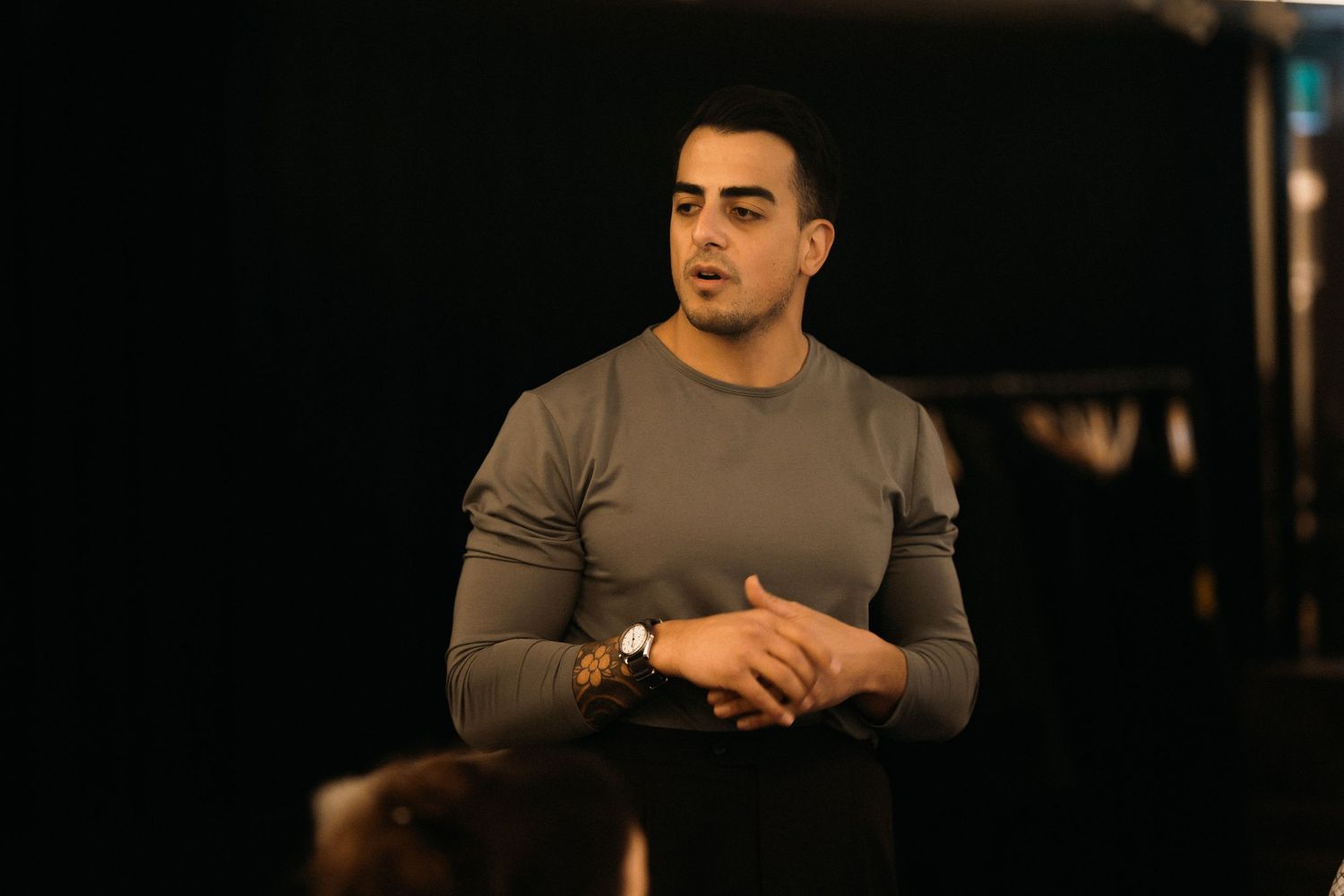 A person wearing a gray long sleeve shirt stands in a dimly lit room with clasped hands.