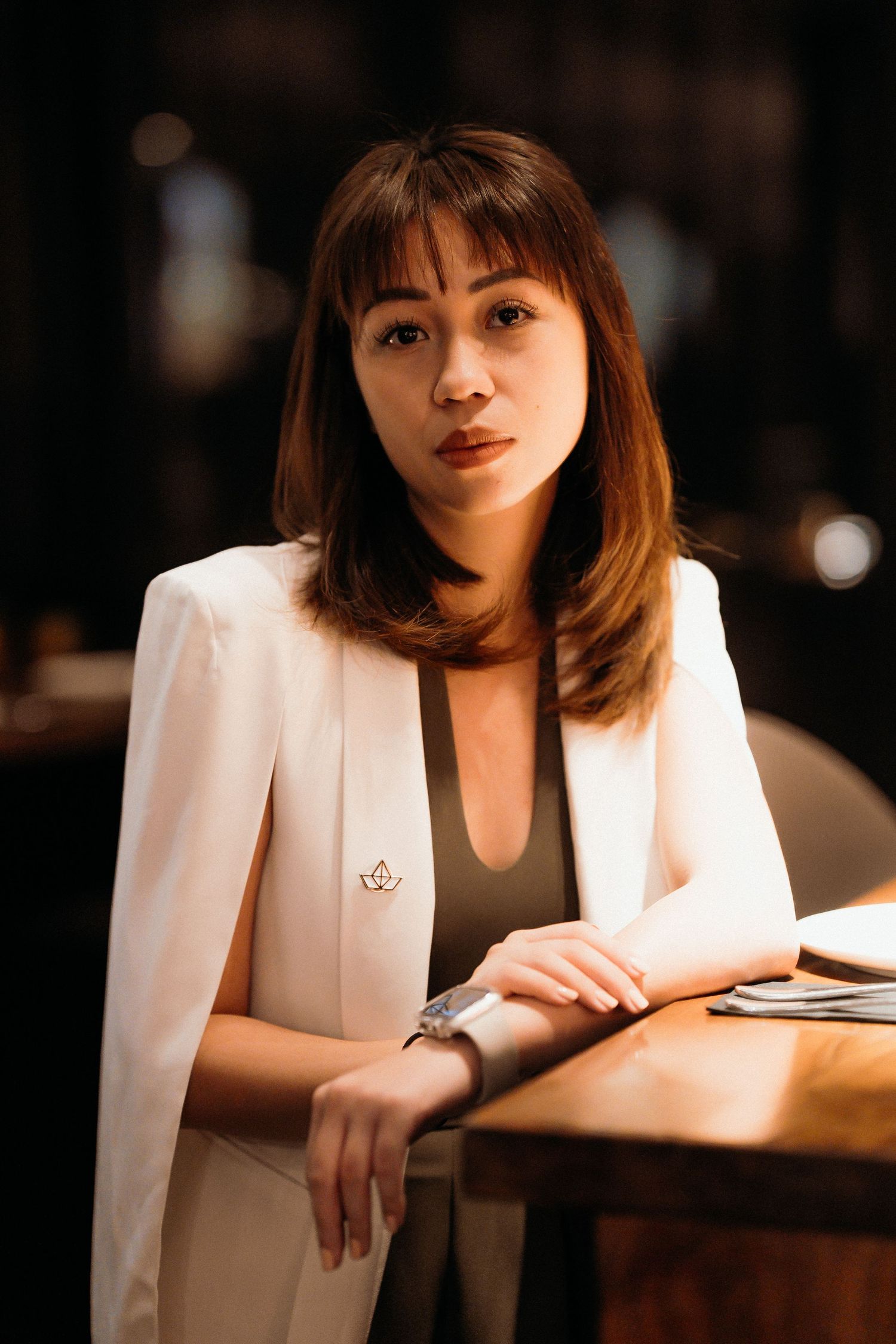 A woman in a white blazer sits at a desk in a dimly lit room with a thoughtful expression.