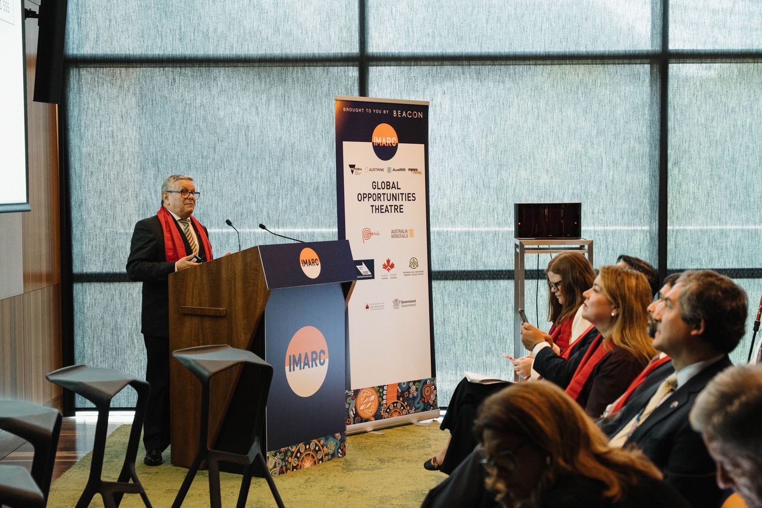 A speaker presents at a professional conference while audience members listen and take notes in a modern meeting room.