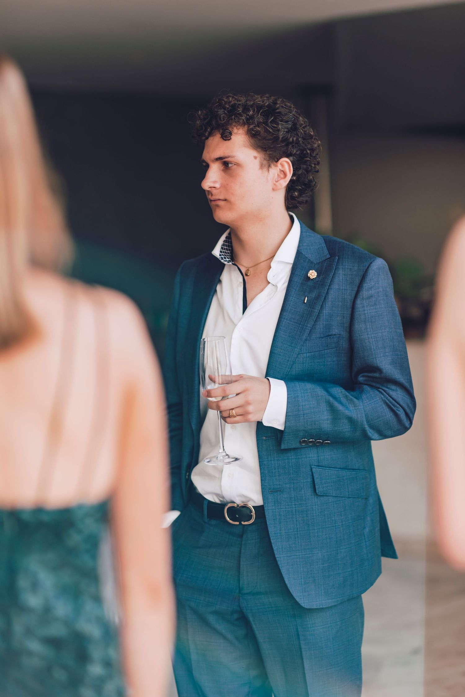 A person in a tailored blue suit holds a wine glass while standing at an elegant event.