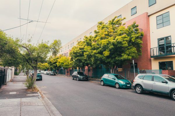 Tree-lined residential street with parked cars and colorful apartment buildings on a sunny day in an urban neighborhood.