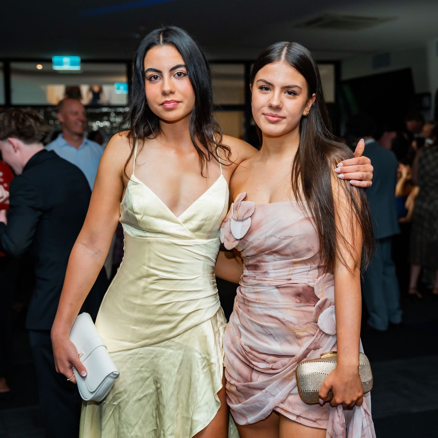 Two women in satin dresses pose together at a formal event, one wearing pale yellow and one in pink.
