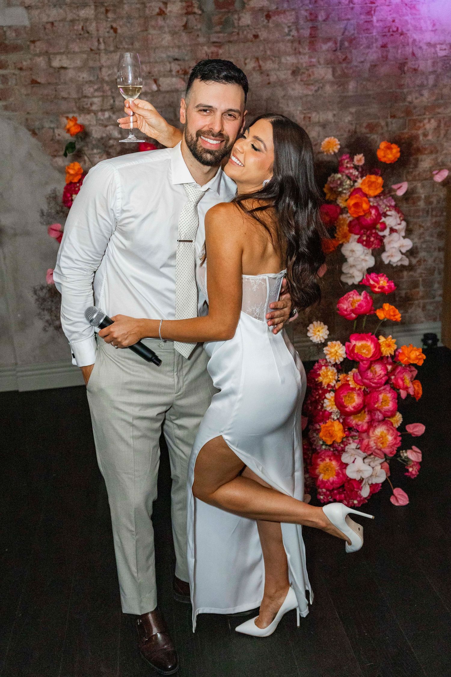 Couple in elegant white attire embrace and celebrate near colorful floral wall arrangement at special event.