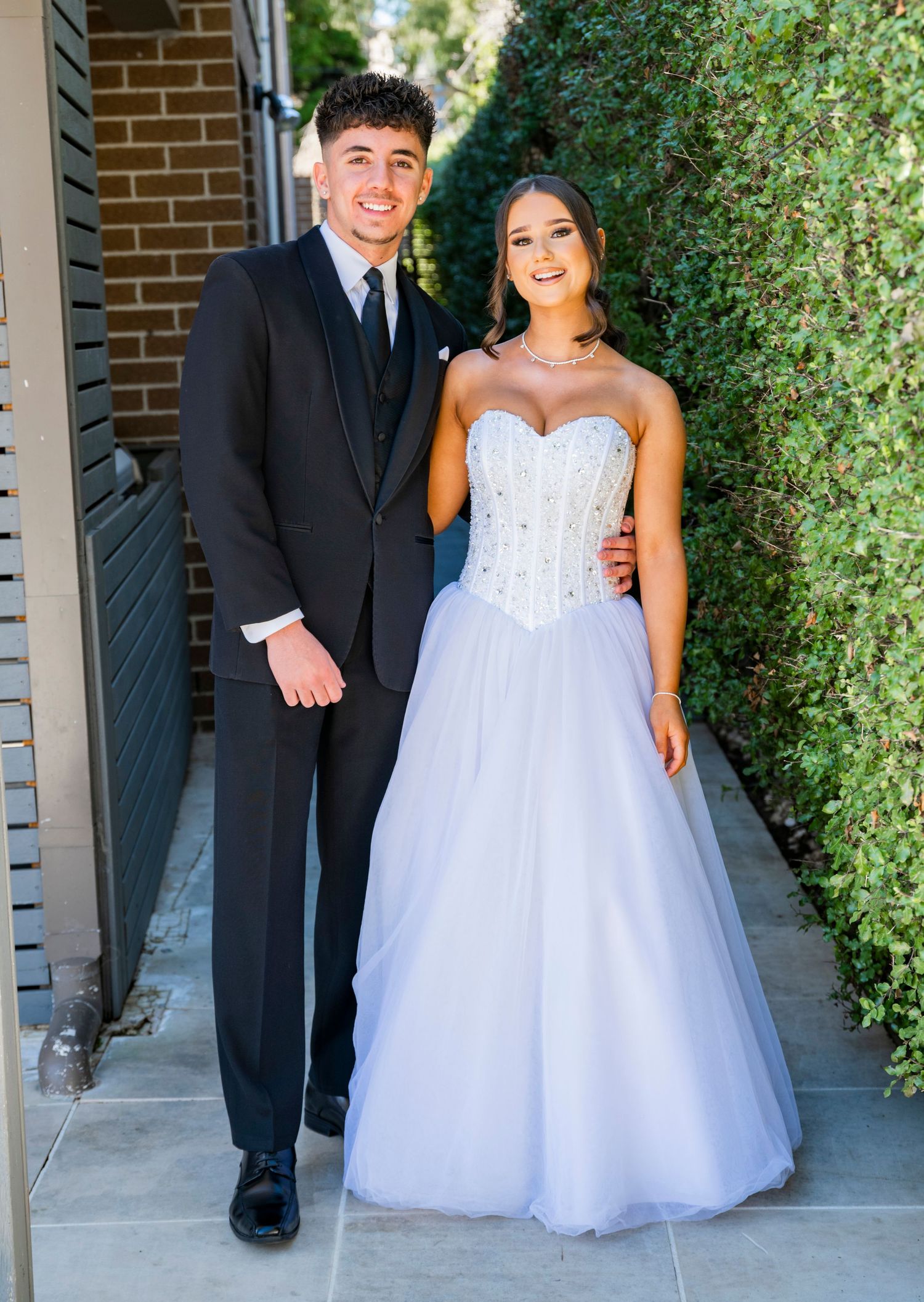 A celebratory wedding scene with people in formal attire including a white ballgown dress and black suit against a brick wall.