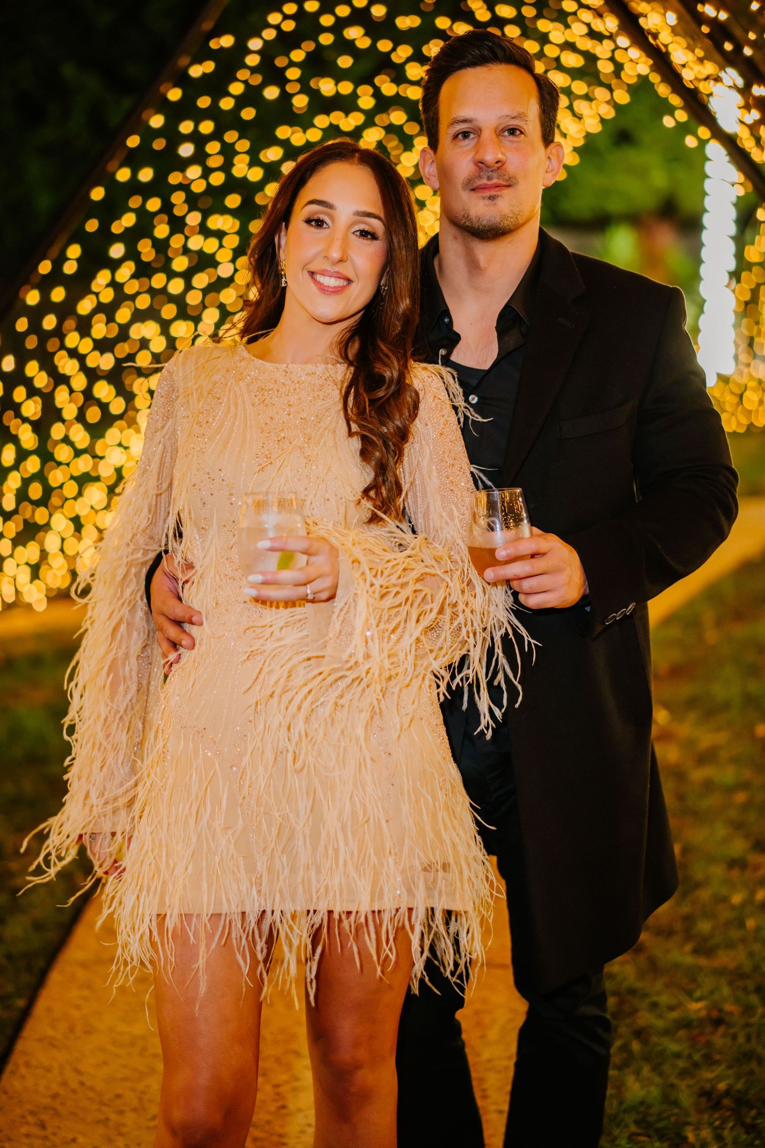 A couple poses outdoors at night with twinkling string lights creating a magical golden bokeh effect in the background.