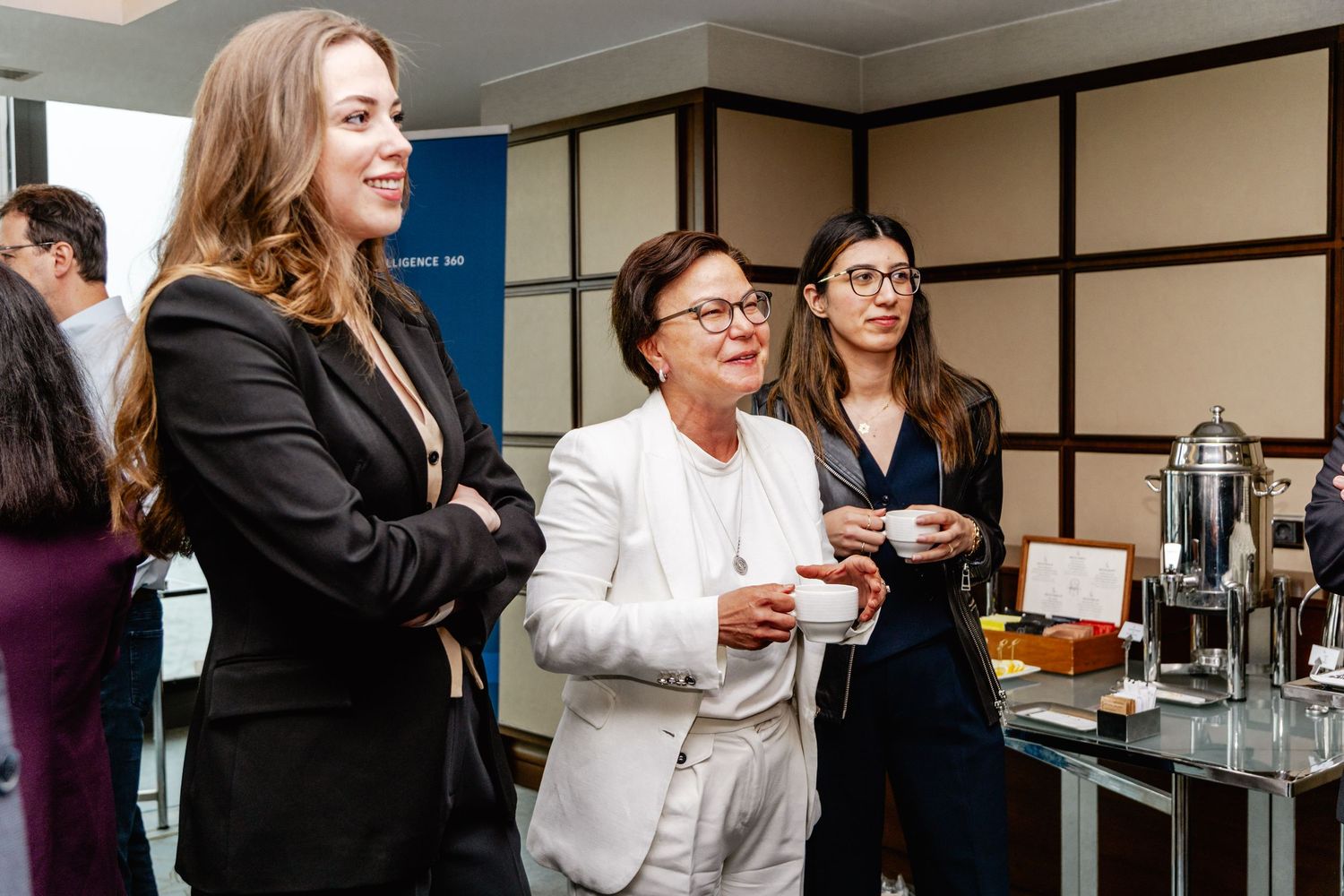 Three professional women networking at a corporate event, standing near a refreshment table in a modern venue.