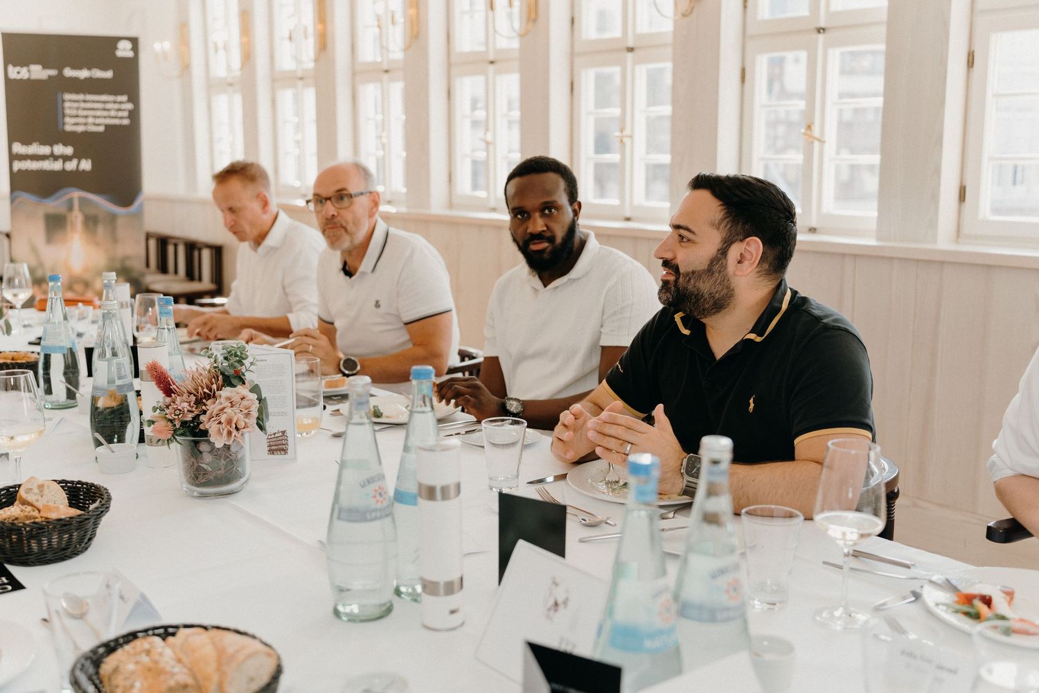Men in white and black polo shirts sit at a conference table with water bottles during a business meeting.