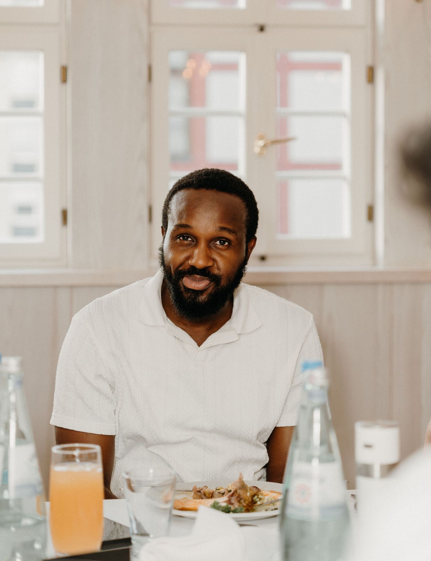 Smiling man in white shirt sitting at a dining table with food and drinks in a bright restaurant.