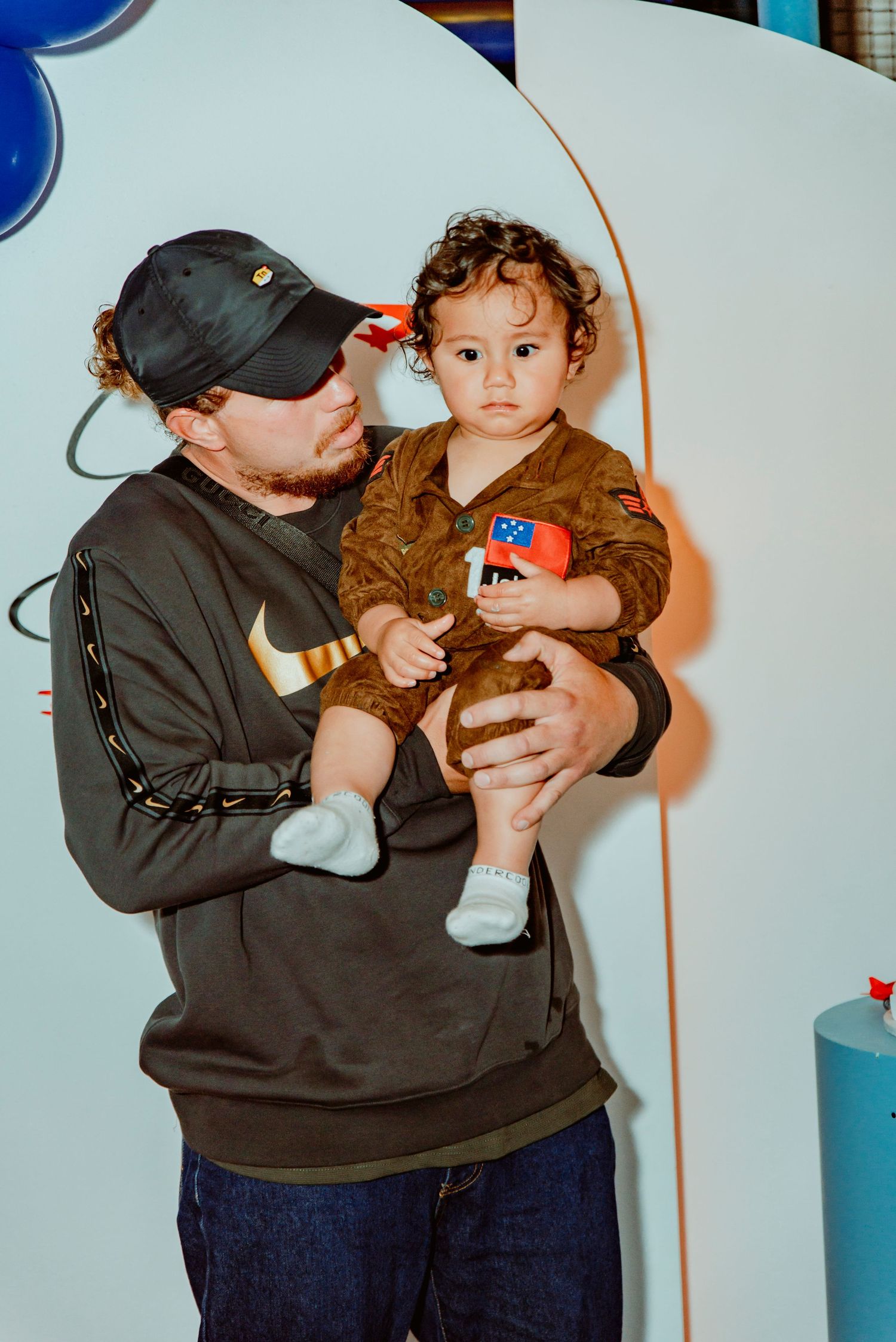 A person in a black baseball cap and sweatshirt holds a toddler wearing a brown outfit and blue socks indoors.