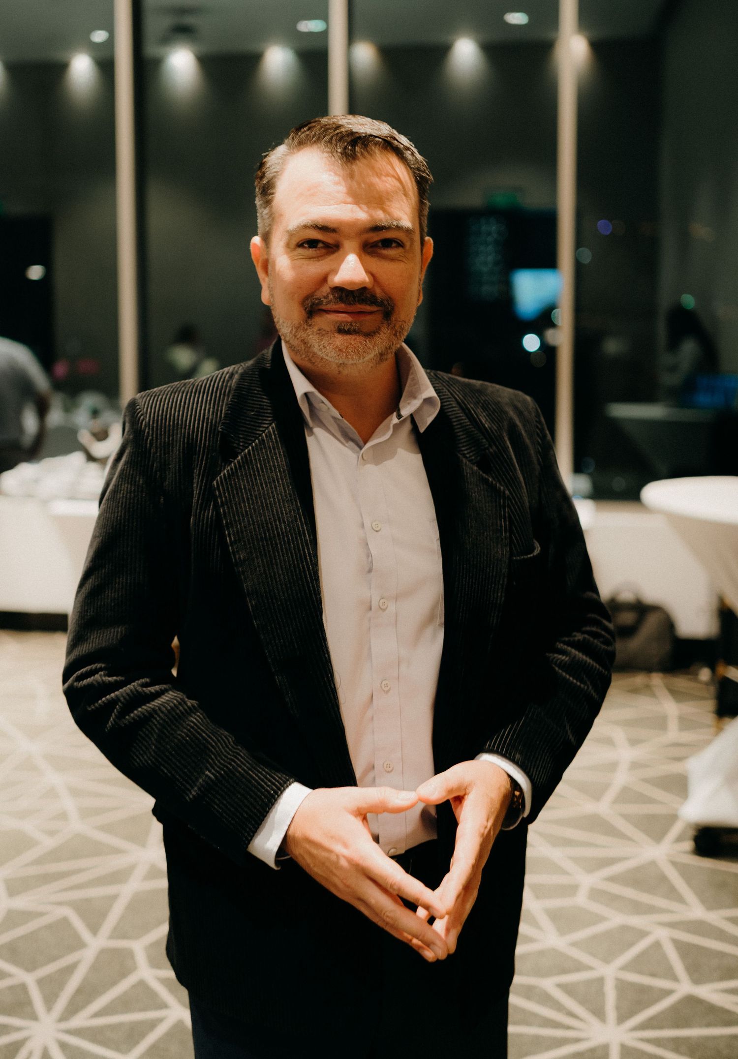 A person in a black suit and white shirt sits in an elegant event space with geometric patterned carpet.
