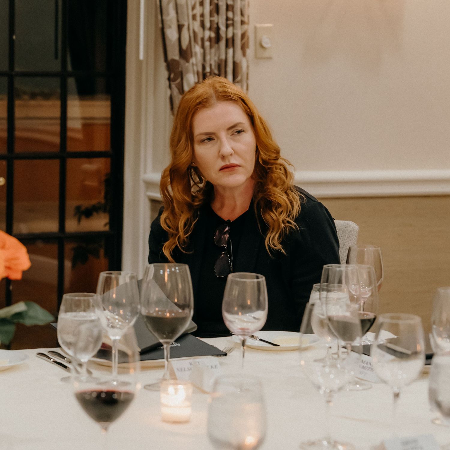 A person sits at an elegant dinner table with wine glasses and candlelight in a moody, intimate setting.