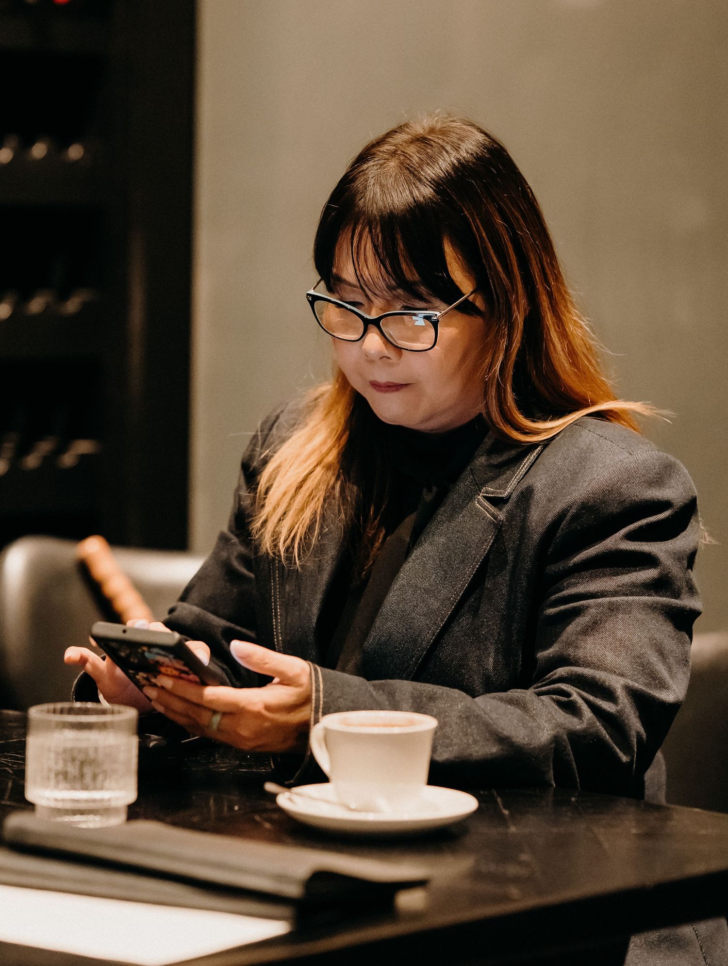 Person in dark blazer sits at restaurant table looking at smartphone while dining alone.