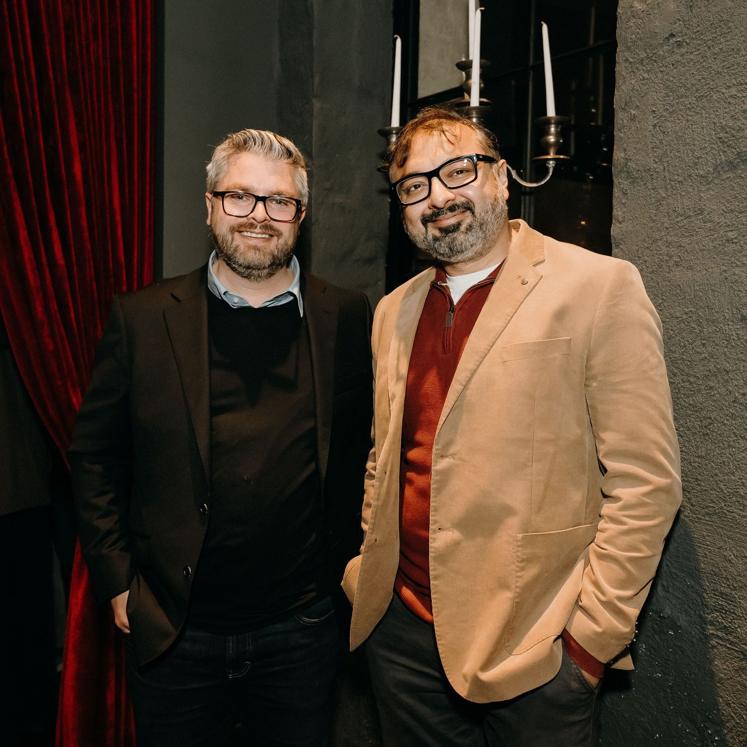 Two men in glasses stand together in front of a dark background with red curtain and candelabra.