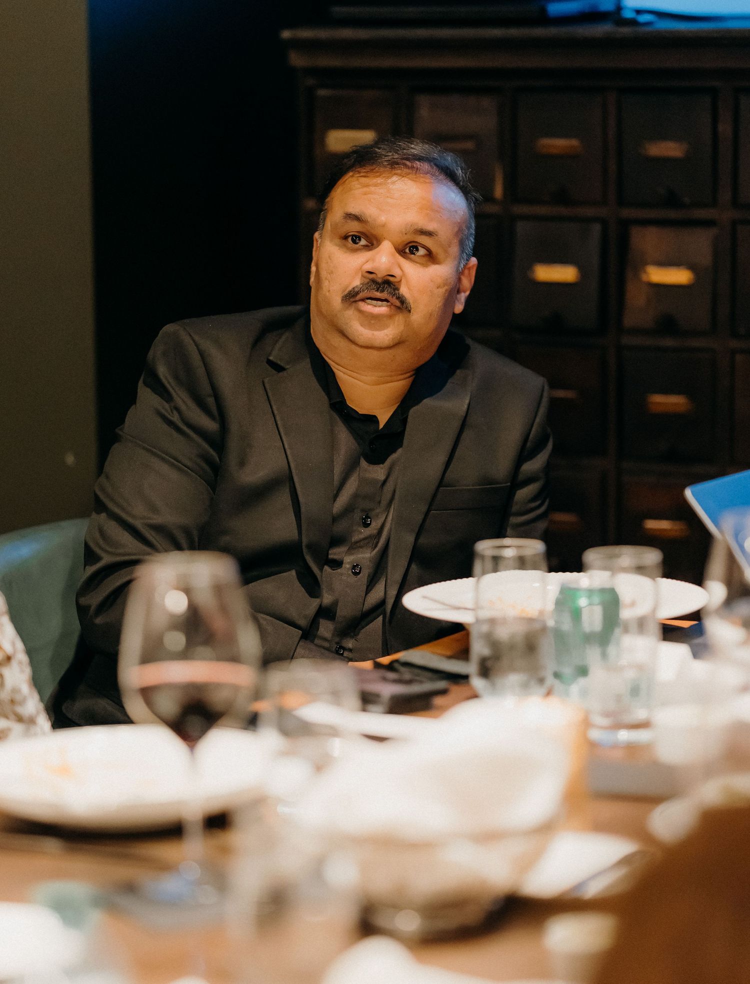 A person in a dark suit sits at a dining table with place settings and glassware in a dimly lit restaurant.
