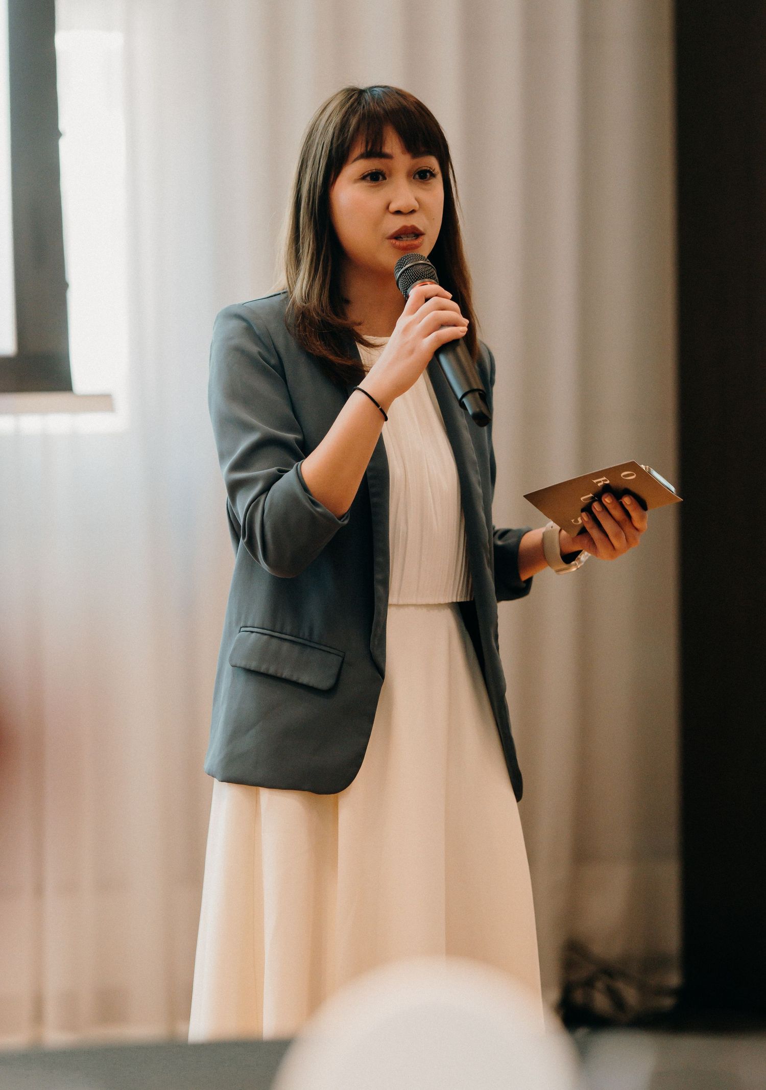 Woman in gray blazer and white dress checks phone while standing against curtained window in office setting.