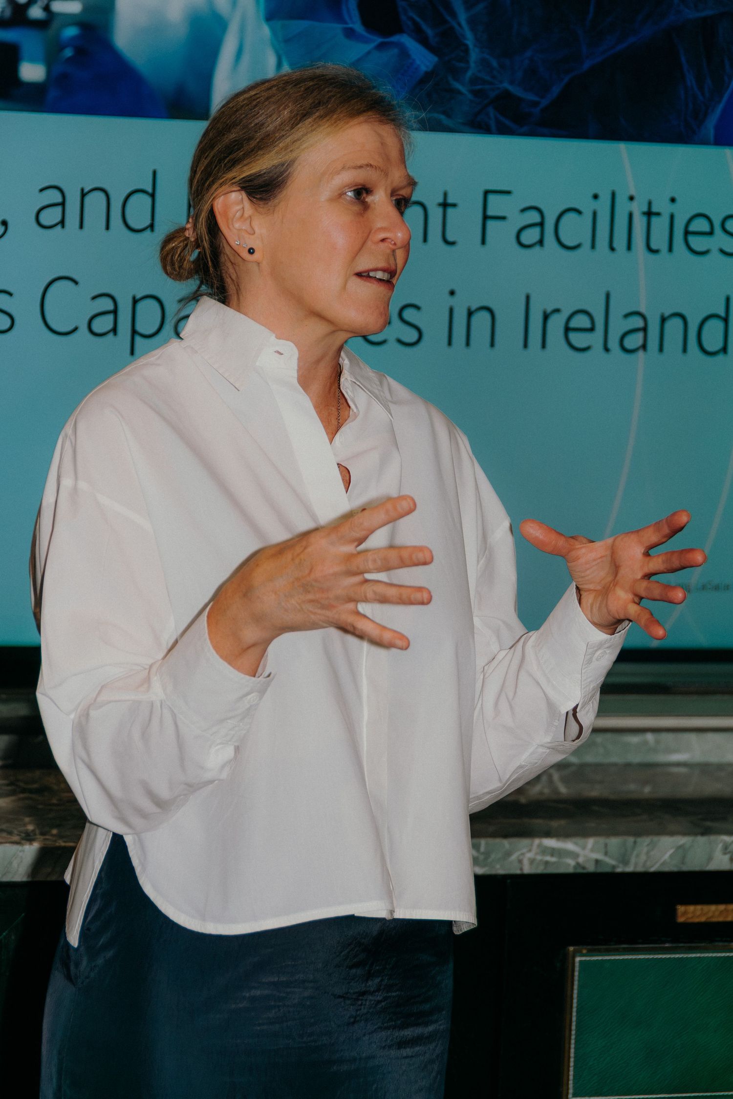 Person in white long sleeve top and dark pants gestures expressively while speaking at a business event.