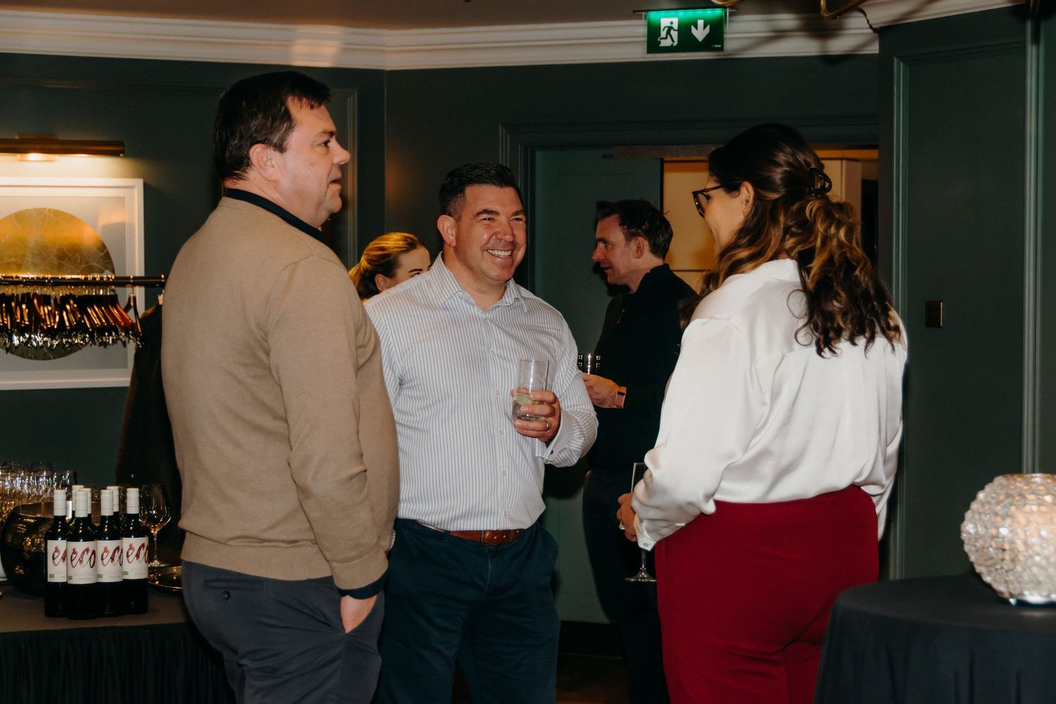 People socializing and having drinks at an indoor gathering in a dark green room with bar decor.