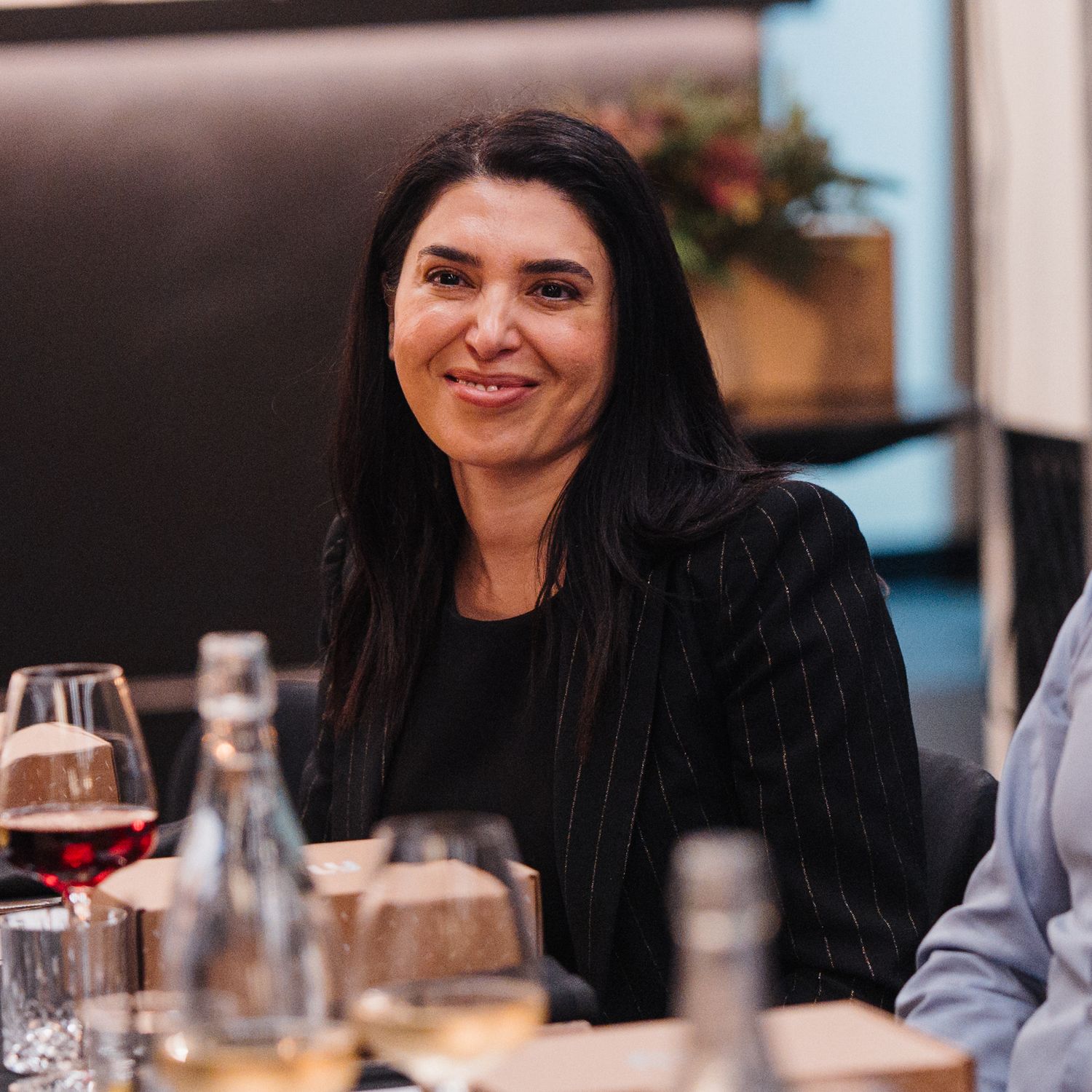 A person with long dark hair smiling while dining at a restaurant with wine glasses and table settings visible.