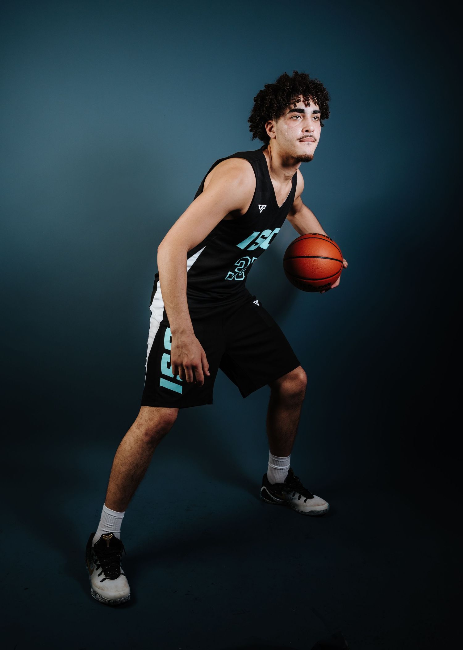 Basketball player in black jersey crouches low with ball in dramatic lighting against dark background.