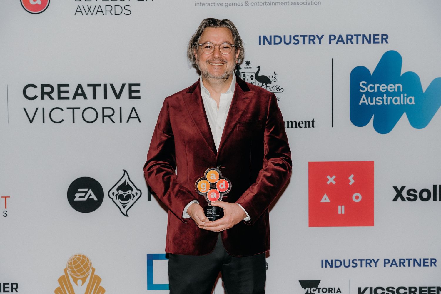A person in a burgundy velvet blazer poses on a red carpet with various sponsor logos and banners in the background.