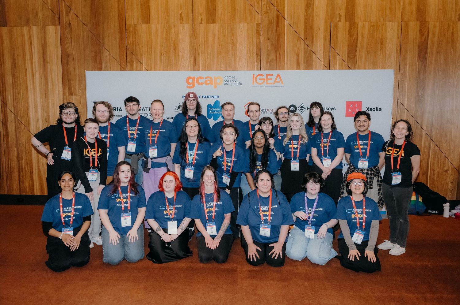 A group of people wearing matching blue shirts sit together against a wooden wall during an event.