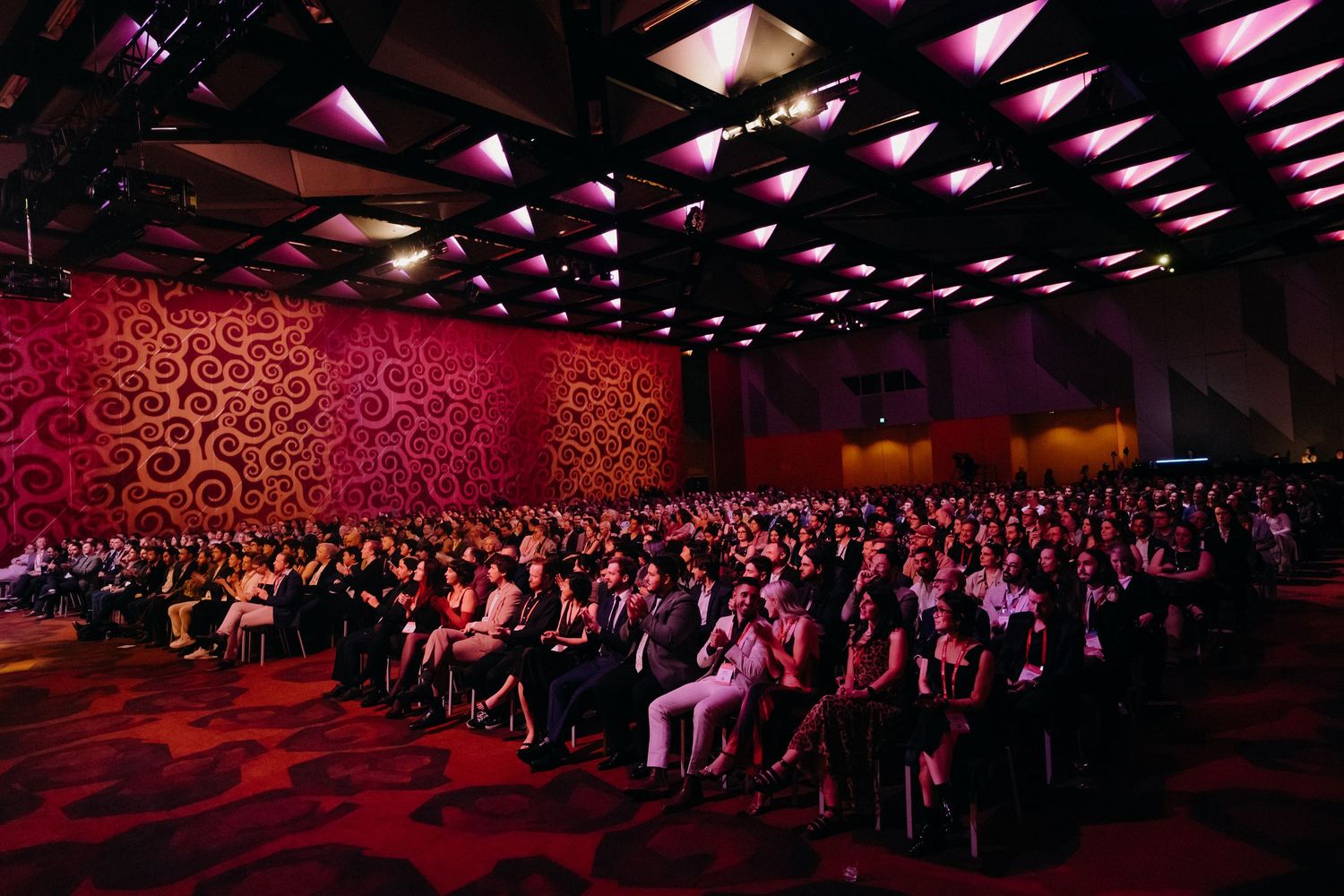 Large audience seated in a dimly lit auditorium with red decorative wall panels and geometric lighting on the ceiling.