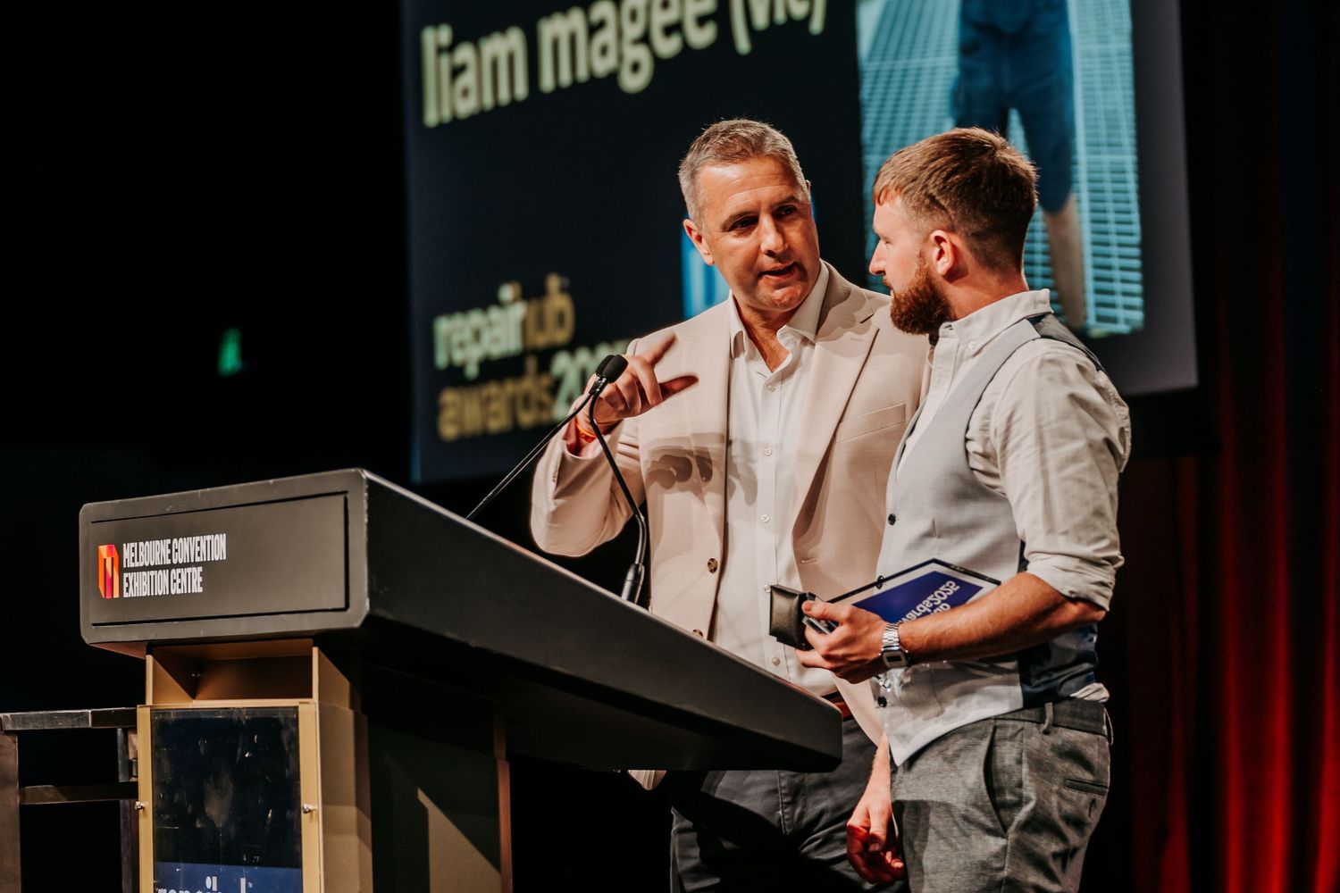 Two speakers engage in animated discussion at a podium during a professional conference presentation.