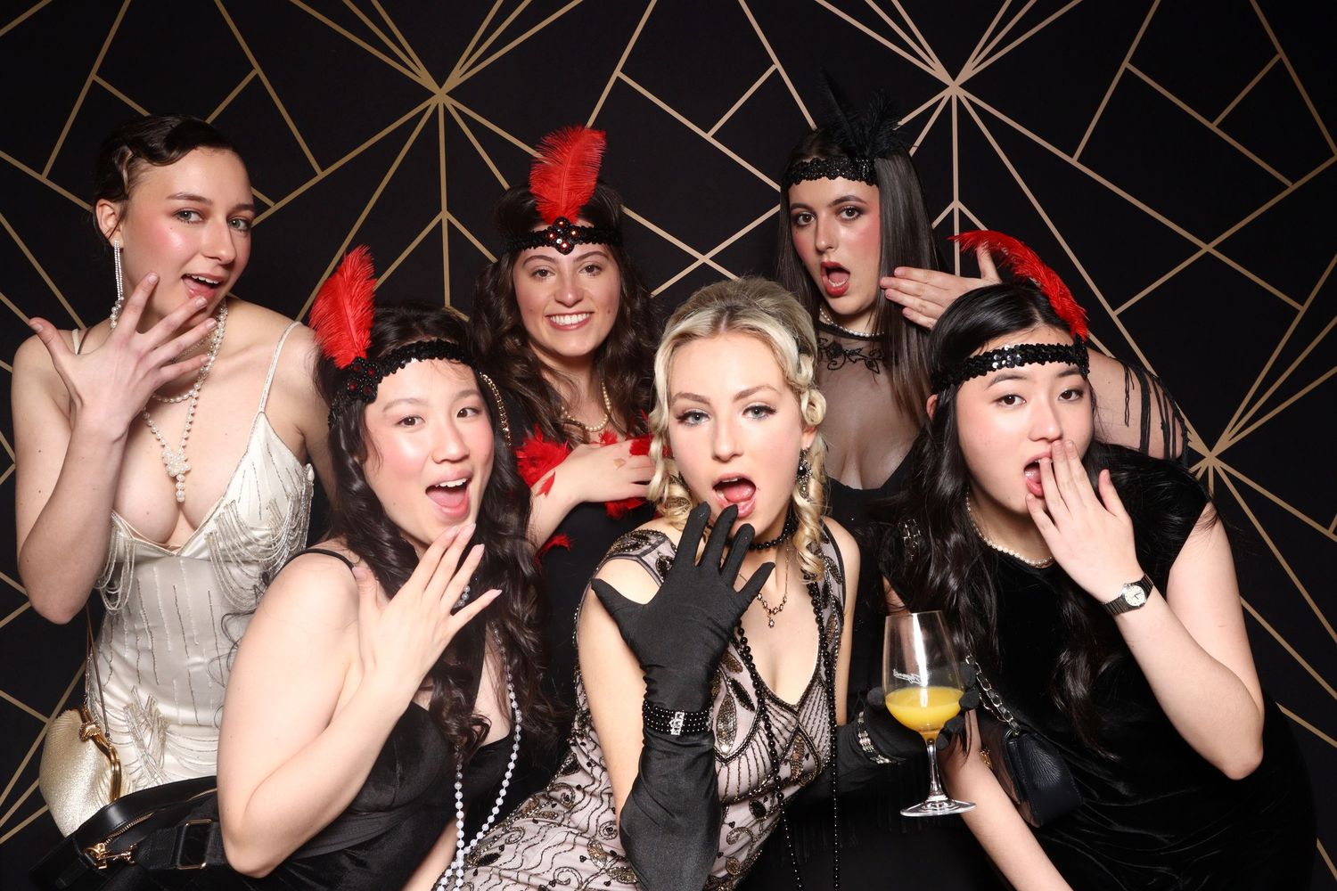 Group of friends in glamorous party attire making surprised expressions while posing at a celebration with geometric backdrop.