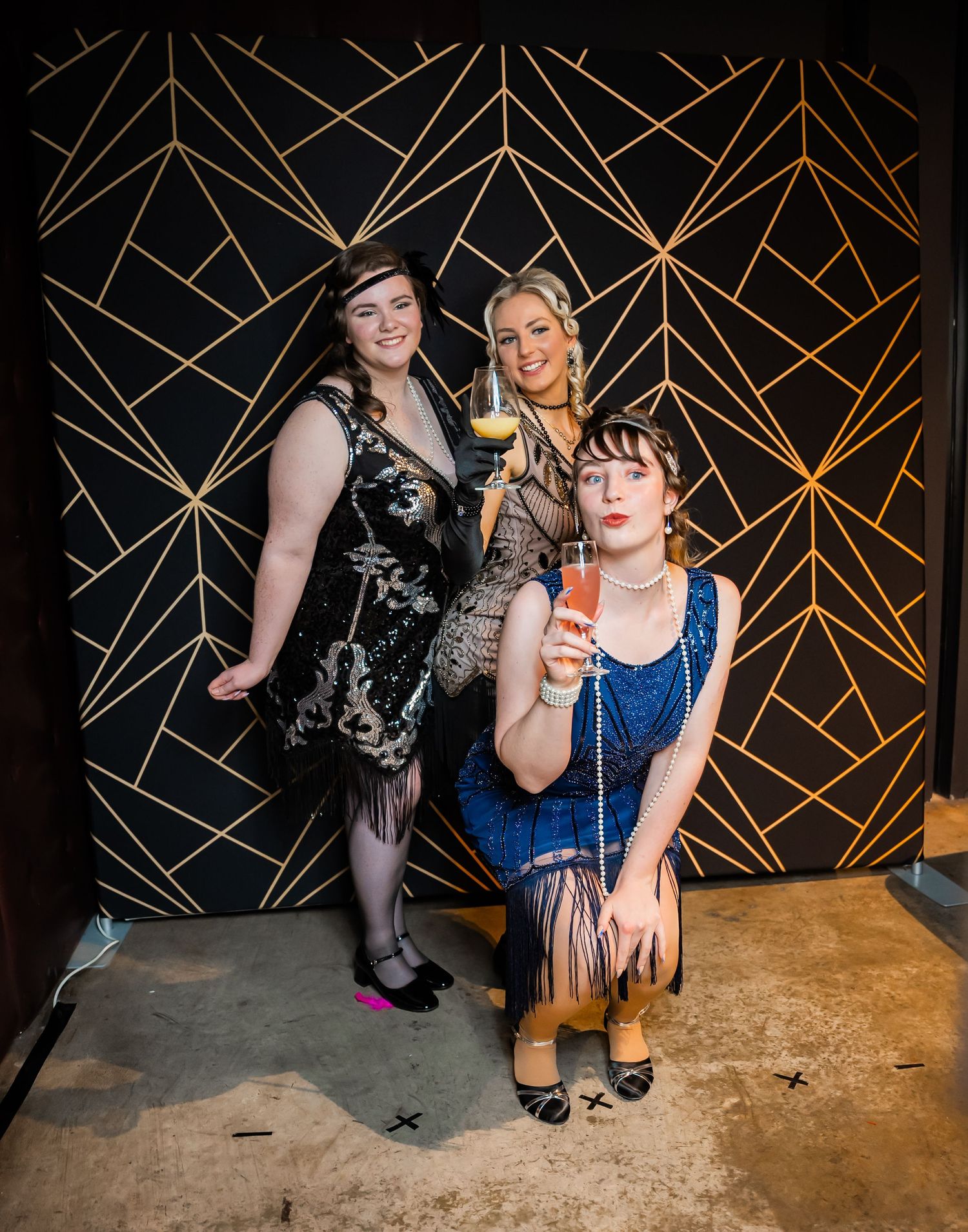 Three people in vintage-inspired cocktail attire pose against an art deco geometric wall pattern at a party.