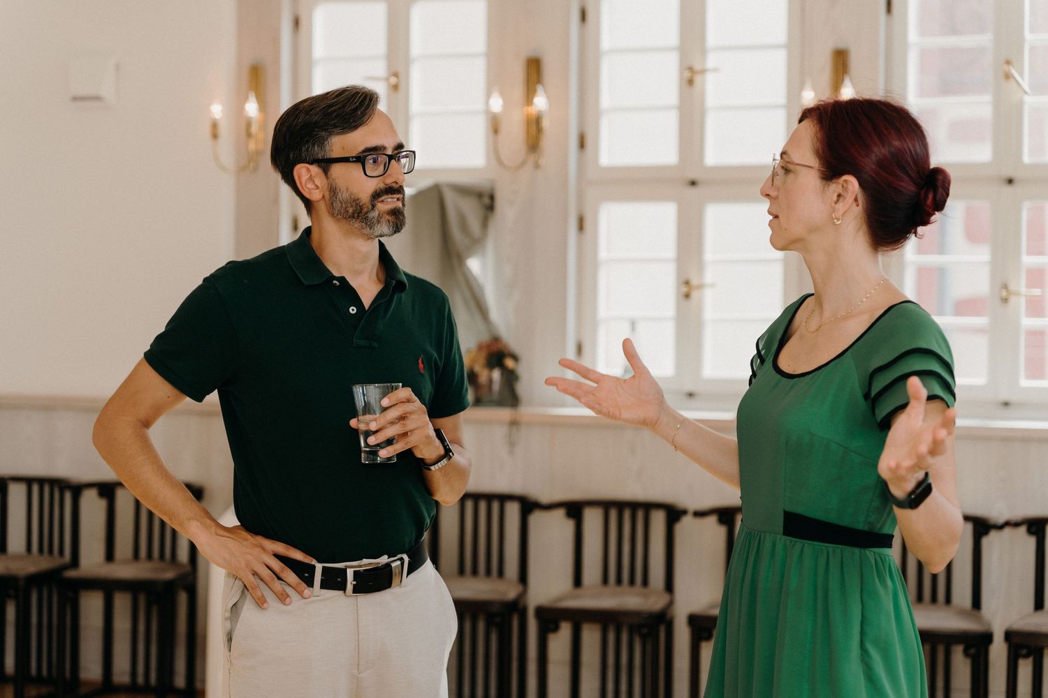 Man and woman in green outfits conversing in elegant hall, man holding drink, woman gesturing expressively.